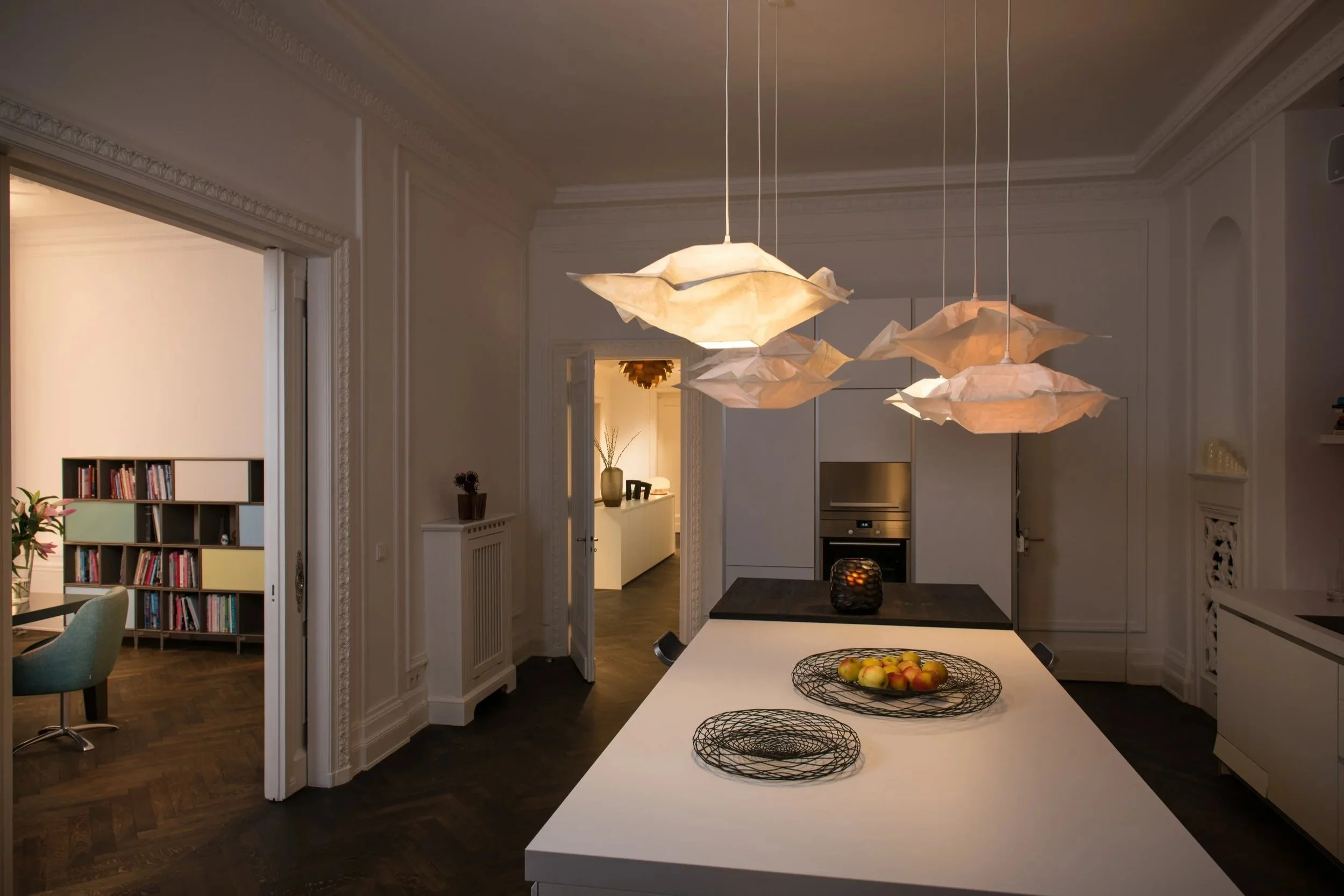 Moderne Küche mit weißen Wänden, dunklem Boden, zentralem Esstisch und dekorativen Papierlampen als Beleuchtung.