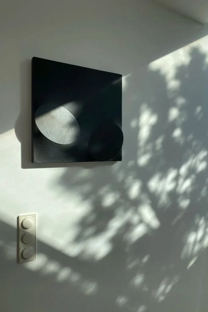 Ein schwarzes Wandbild mit runden Formen, Schatten und Lichtmustern an einer weißen Wand, darunter ein Lichtschalter mit drei runden Knöpfen.