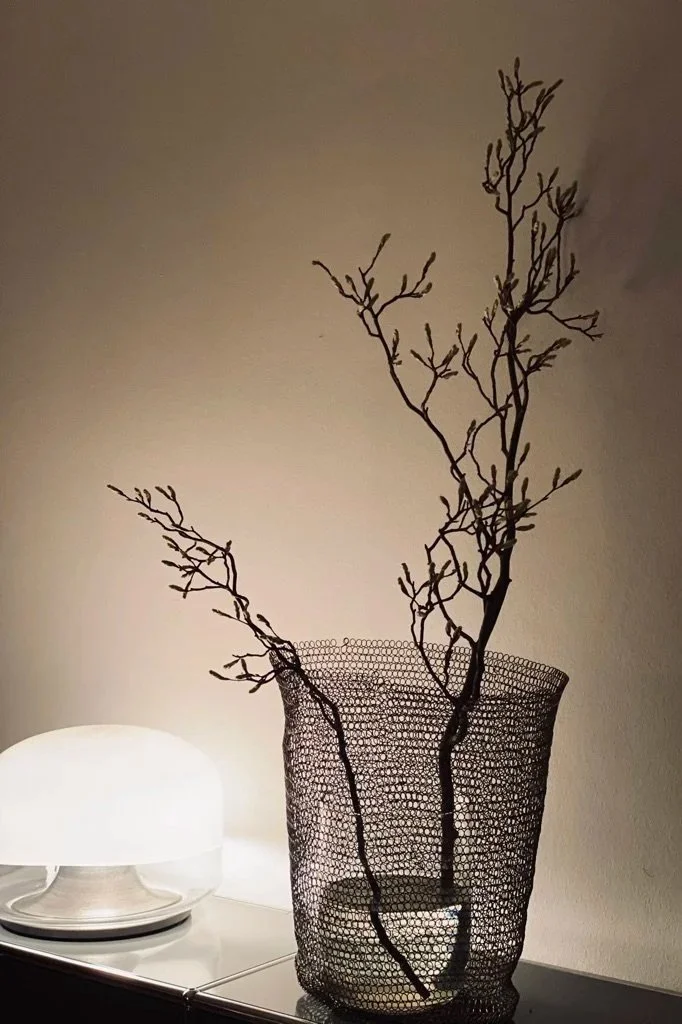 Schön eingerichtete Ecke mit einer beleuchteten weißen Lampe auf einem Nachttisch und einem dekorativen schwarzen Drahtgefäß mit trockenen, verzweigten Zweigen.
