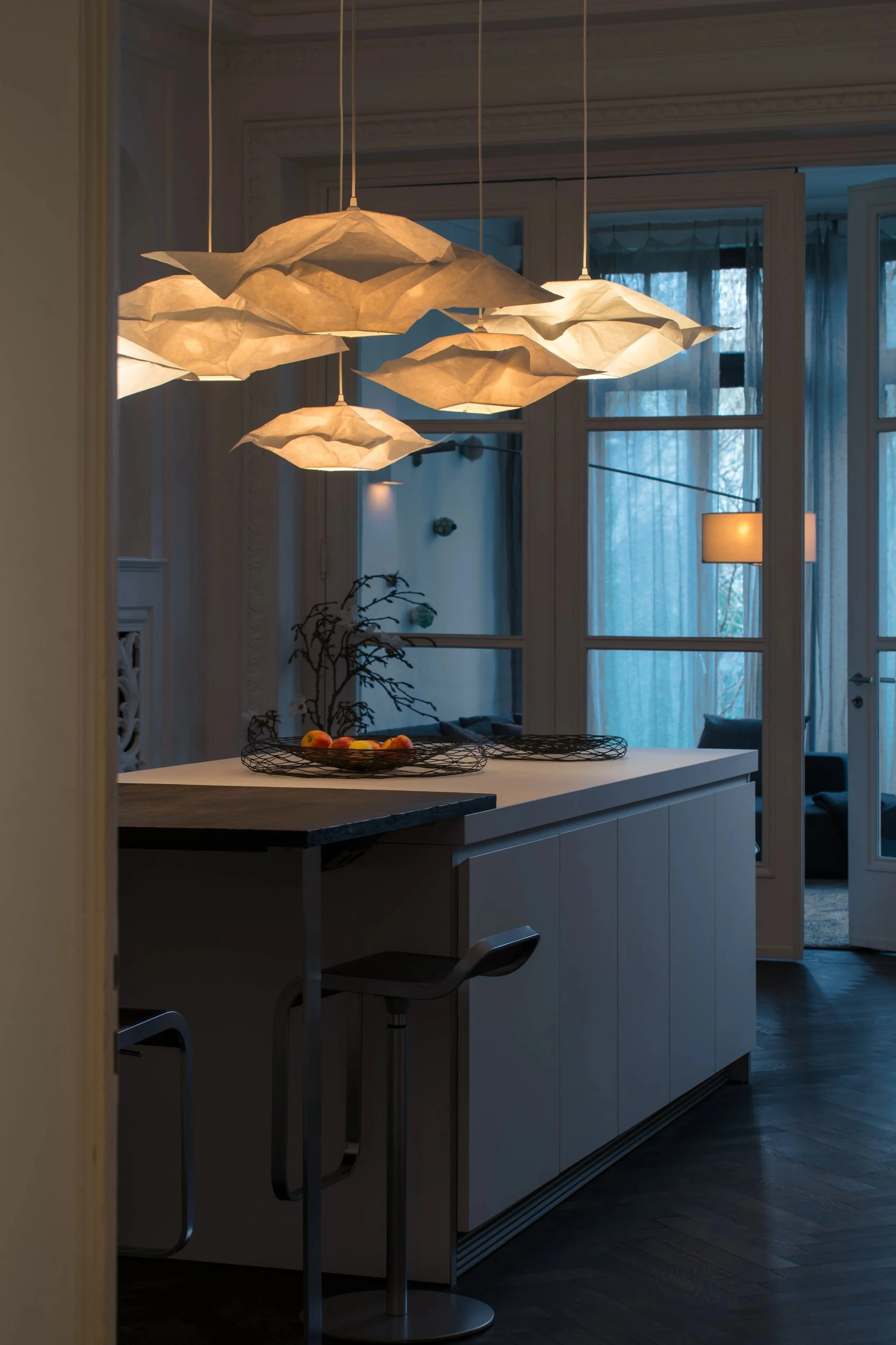 Moderner Essbereich mit hängenden Lampen aus Papier, einer weißen Kücheninsel mit Obstkorb und Blick auf ein Wohnzimmer mit großen Fenstern und Vorhängen im Hintergrund.