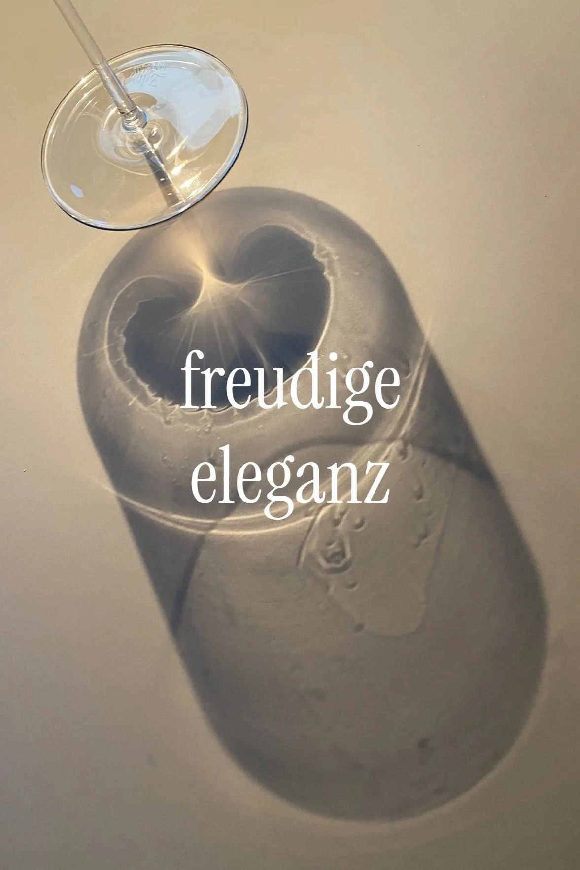 Ein Weinglas auf einer hellen Oberfläche, das einen Schatten mit einer Herzform projiziert. Über dem Schatten steht in weißer Schrift 'freudige eleganz'.