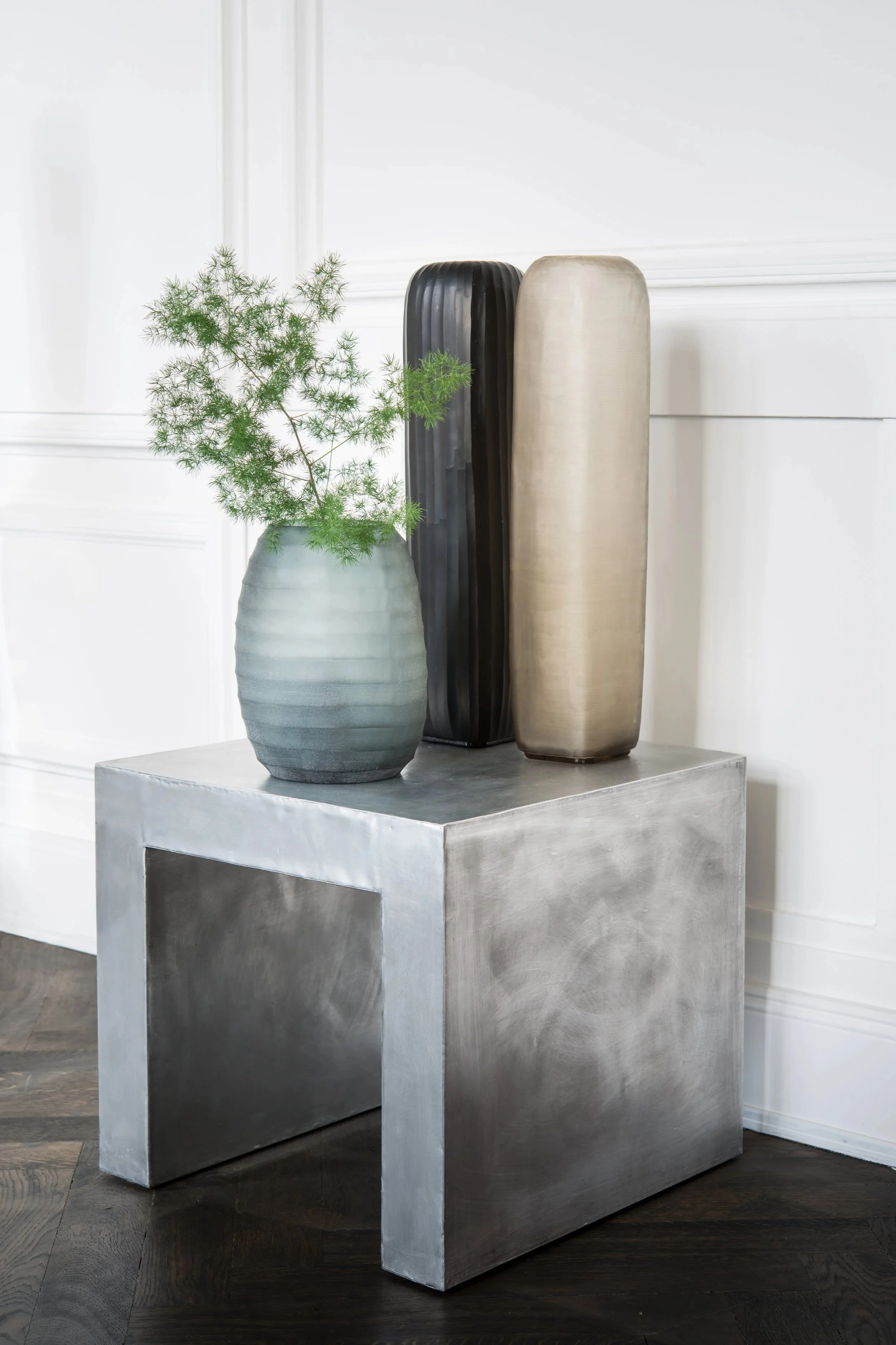 Eine Vase mit grünem Zweig und drei dekorative Objekte auf einem silbernen Tisch vor einer weißen Wand.