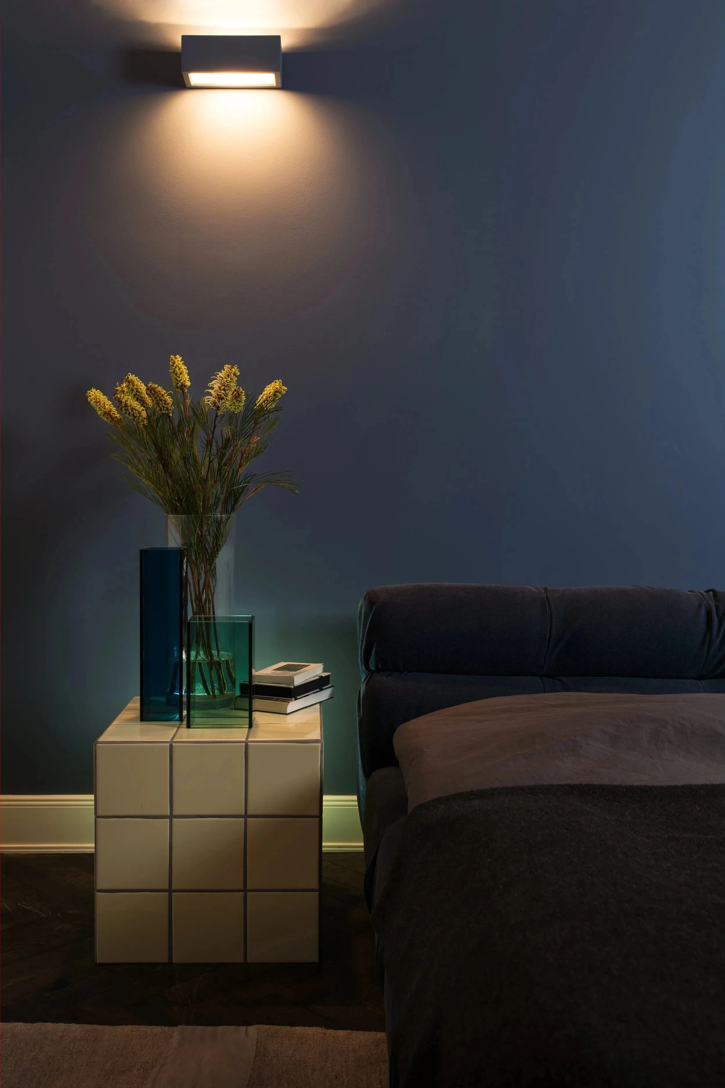 Ein gemütliches Wohnzimmer mit einer dunklen Couch, einem weißen Kachel-Nachtisch, einer Vase mit gelben Blumen, mehreren Büchern und moderner Wandbeleuchtung an einer blauen Wand.