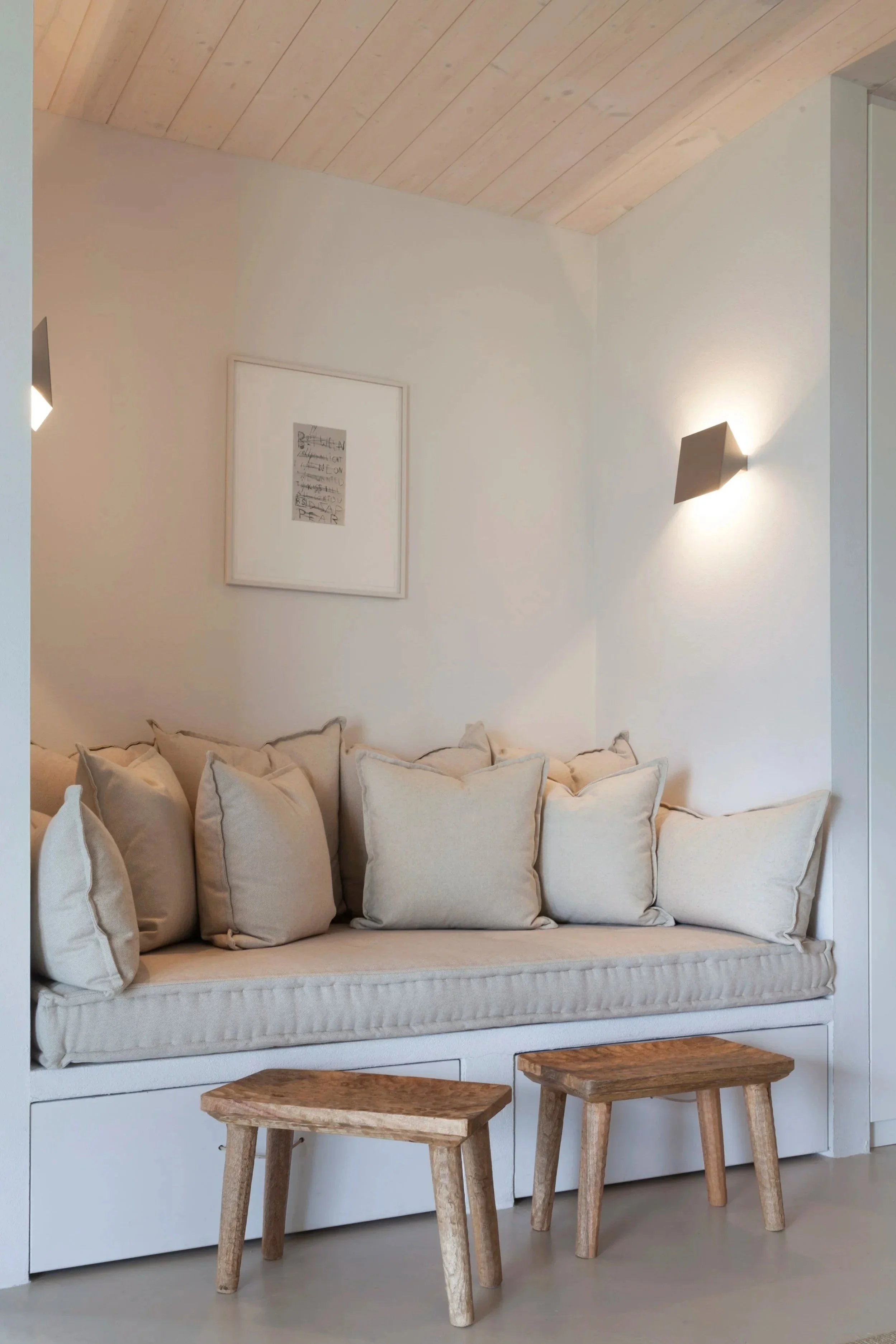 Ein gemütliches Wohnzimmer mit einer weißen Couch, mehreren Kissen, zwei kleinen Holzhockern und einer Wandlampe an einer weißen Wand unter einer Holzdachdecke.