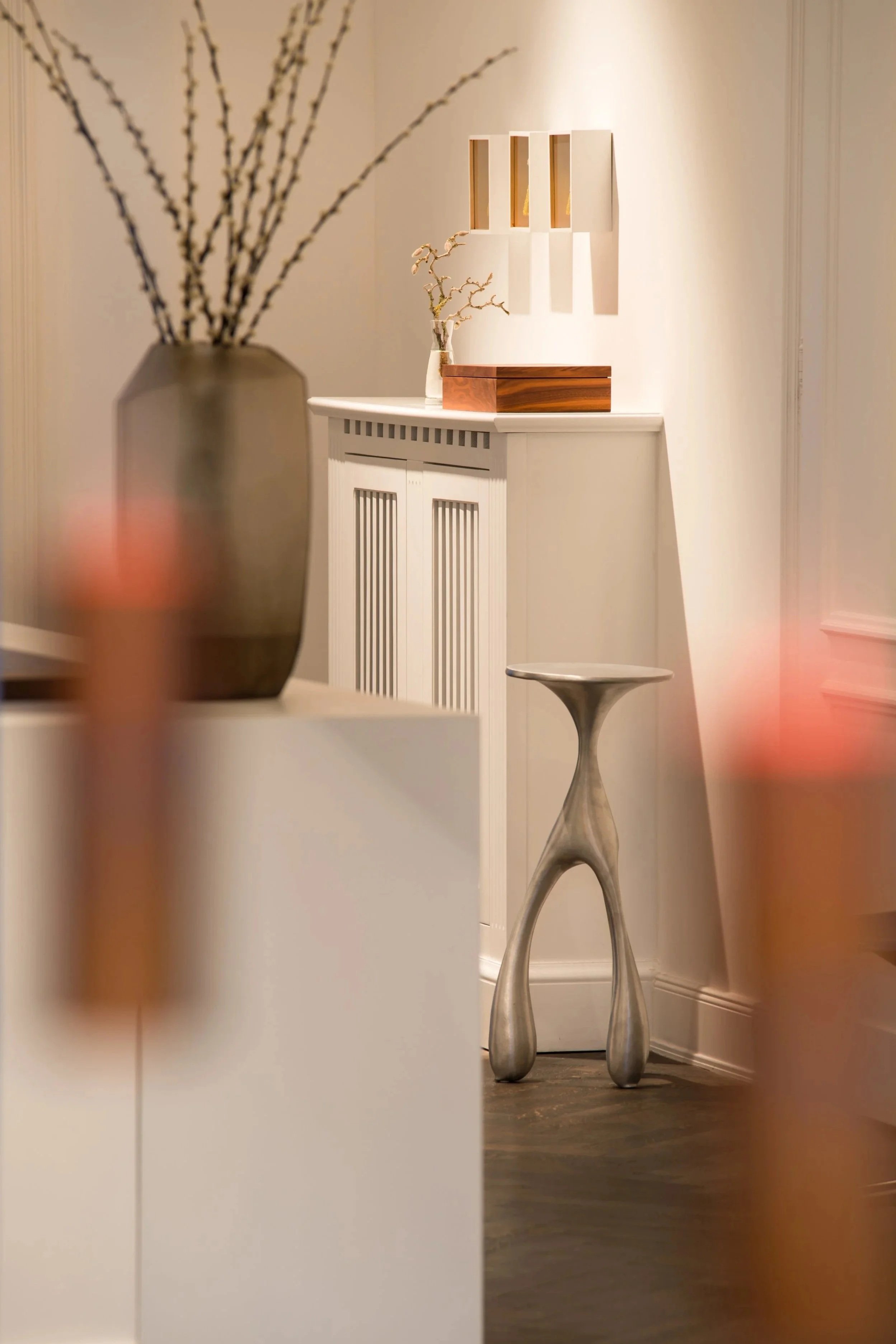 Ein Blick in ein modernes, minimalistisches Interieur mit weißen Wänden und dunklem Holzboden. Im Hintergrund steht ein weißer Schrank mit vertikalen Linien, oben auf einer Ablage steht eine Vase mit Zweigen. Im Vordergrund sind unscharf einige Gegen