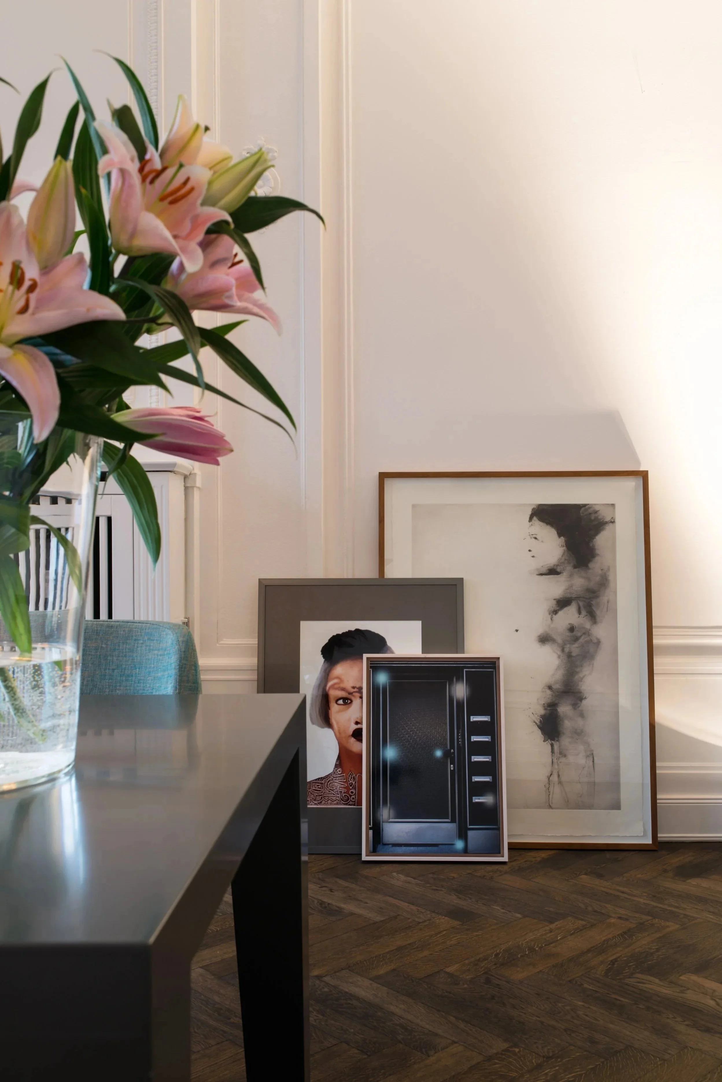 Blumenstrauß mit pinken Lilien auf Tisch, gegen Wand mit mehreren Kunstbildern, dunkler Holzfußboden