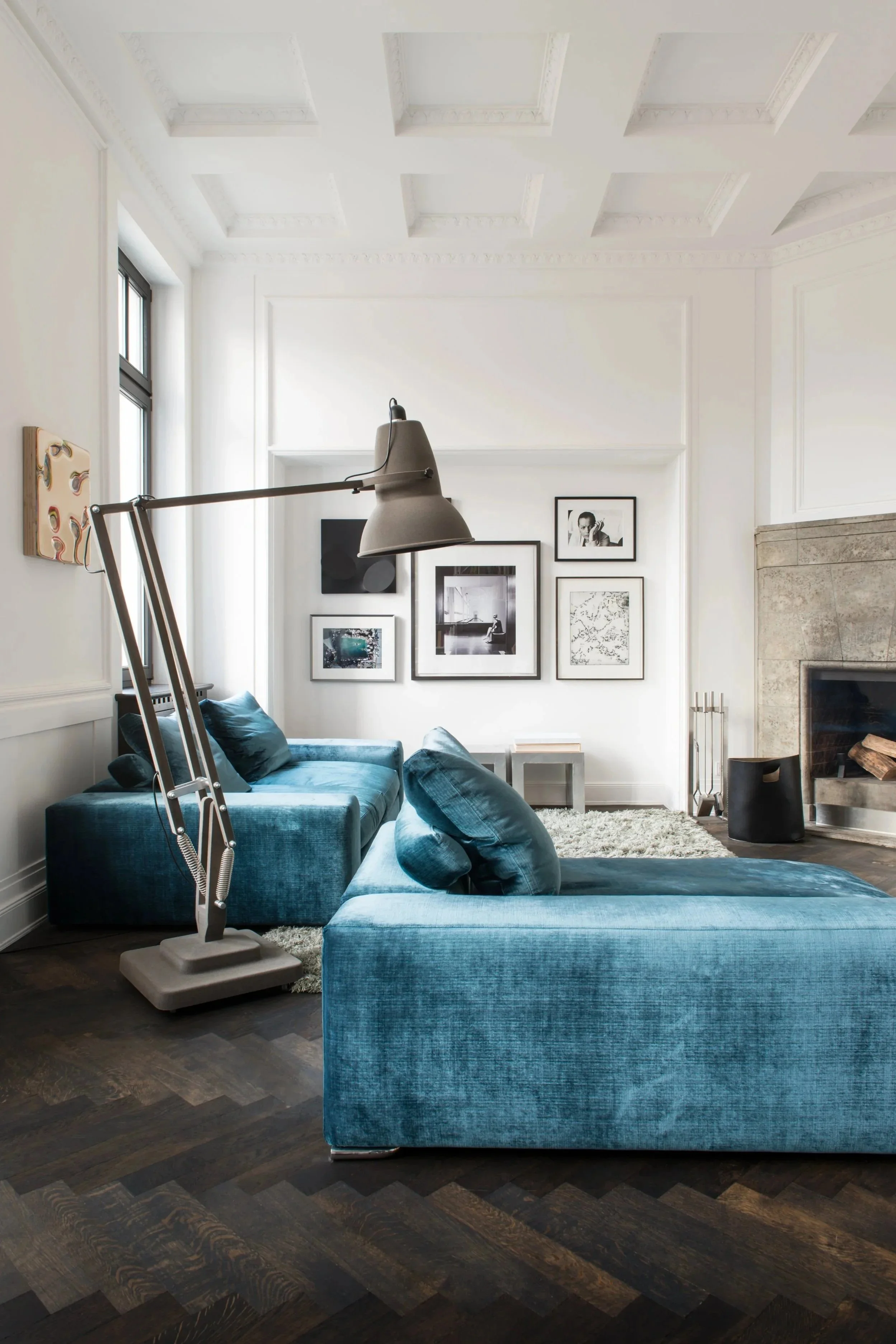 Ein modernes Wohnzimmer mit weißen Wänden, hoher Decke, dunklem Holzboden und zwei blaue Samtsofas. Es gibt eine Stehlampe über den Sofas, eine Feuerstelle aus Stein an der Wand, und eine Bildergalerie mit schwarzen und weißen Fotografien. Ein Fenste