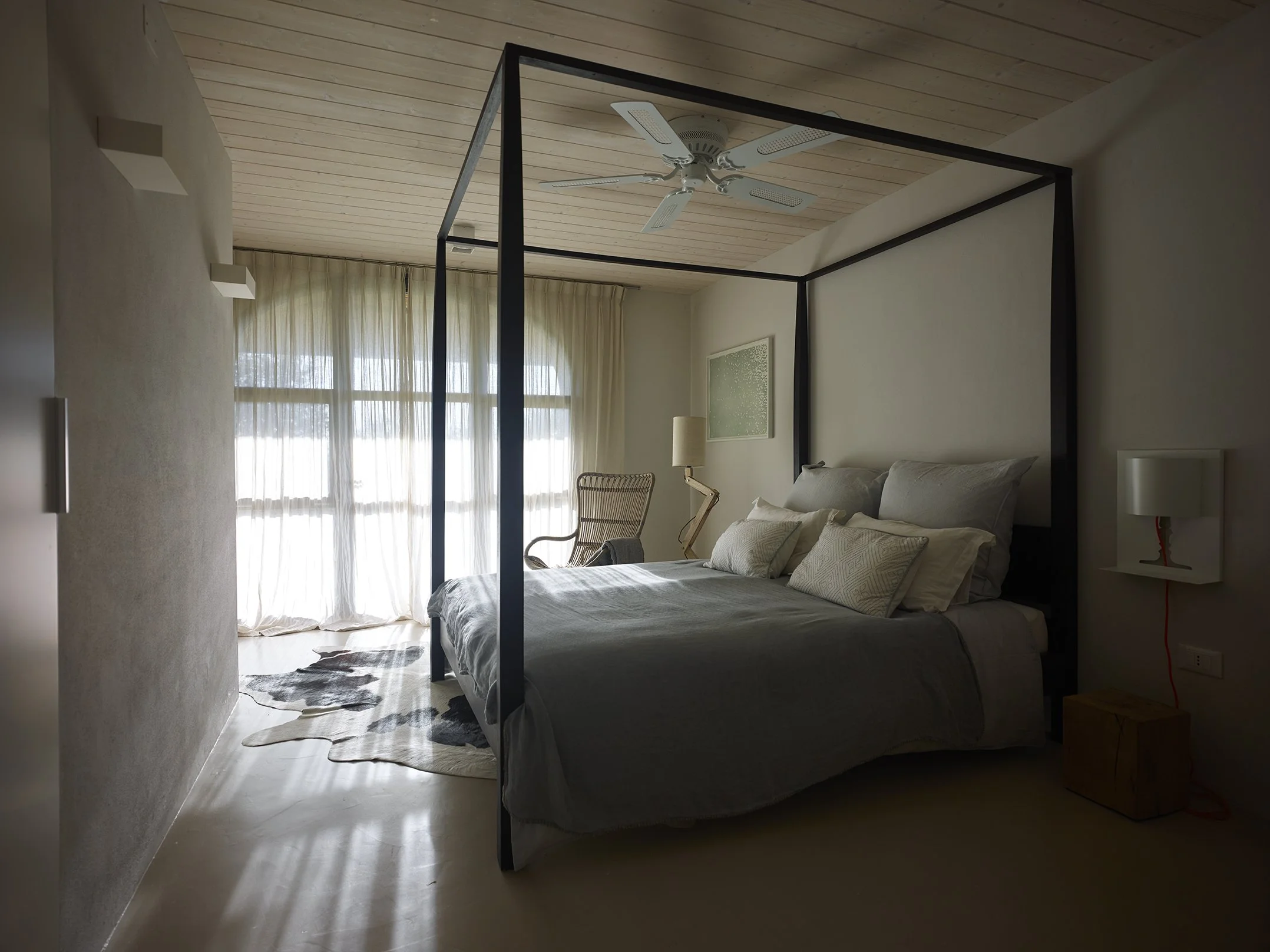 Ein Schlafzimmer mit einem Himmelbett, einer Stehlampe, einem Schreibtischstuhl, einem Wandbild und großen Fenstern mit weißen Vorhängen.