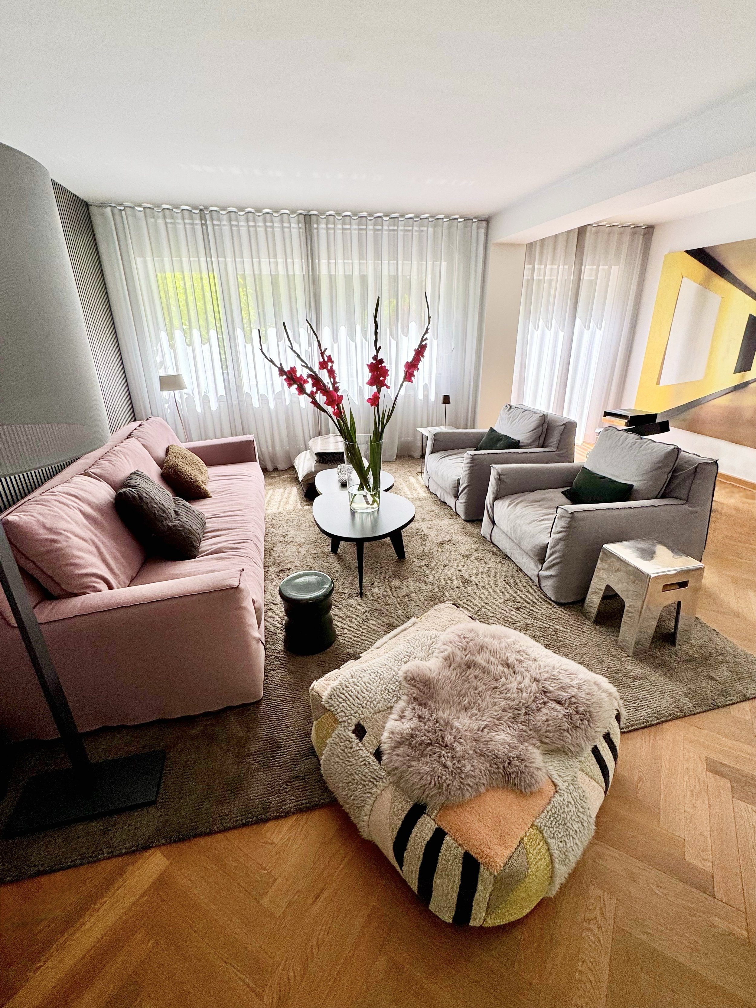 Hell erleuchtetes Wohnzimmer mit einem pinkfarbenen Sofa, weißen Sessel, einer Couchtisch mit Blumenvasen, einem kleinen schwarzen Hocker, einem Teppich und einem gemusterten Fußhocker mit Fell und Decke.