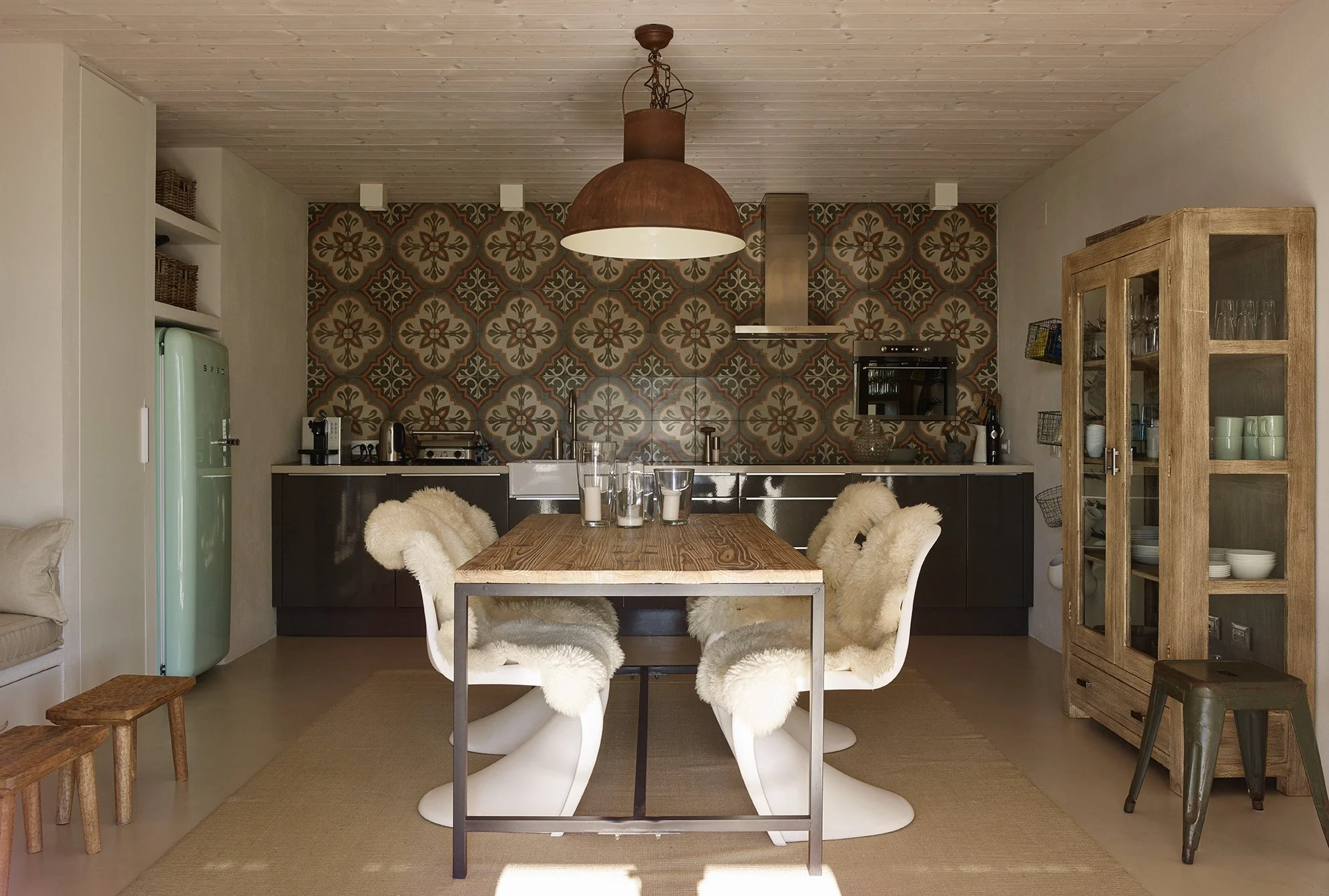 Moderner Küchenraum mit Essbereich, Holztisch, vier Stühlen mit Fellbezügen, mit Kerzen, in einer Mischung aus rustikalem und skandinavischem Stil, mit darunter liegendem Teppich, einer grünen Retro-Kühlschrank, einer offenen Vitrine mit Geschirr, un