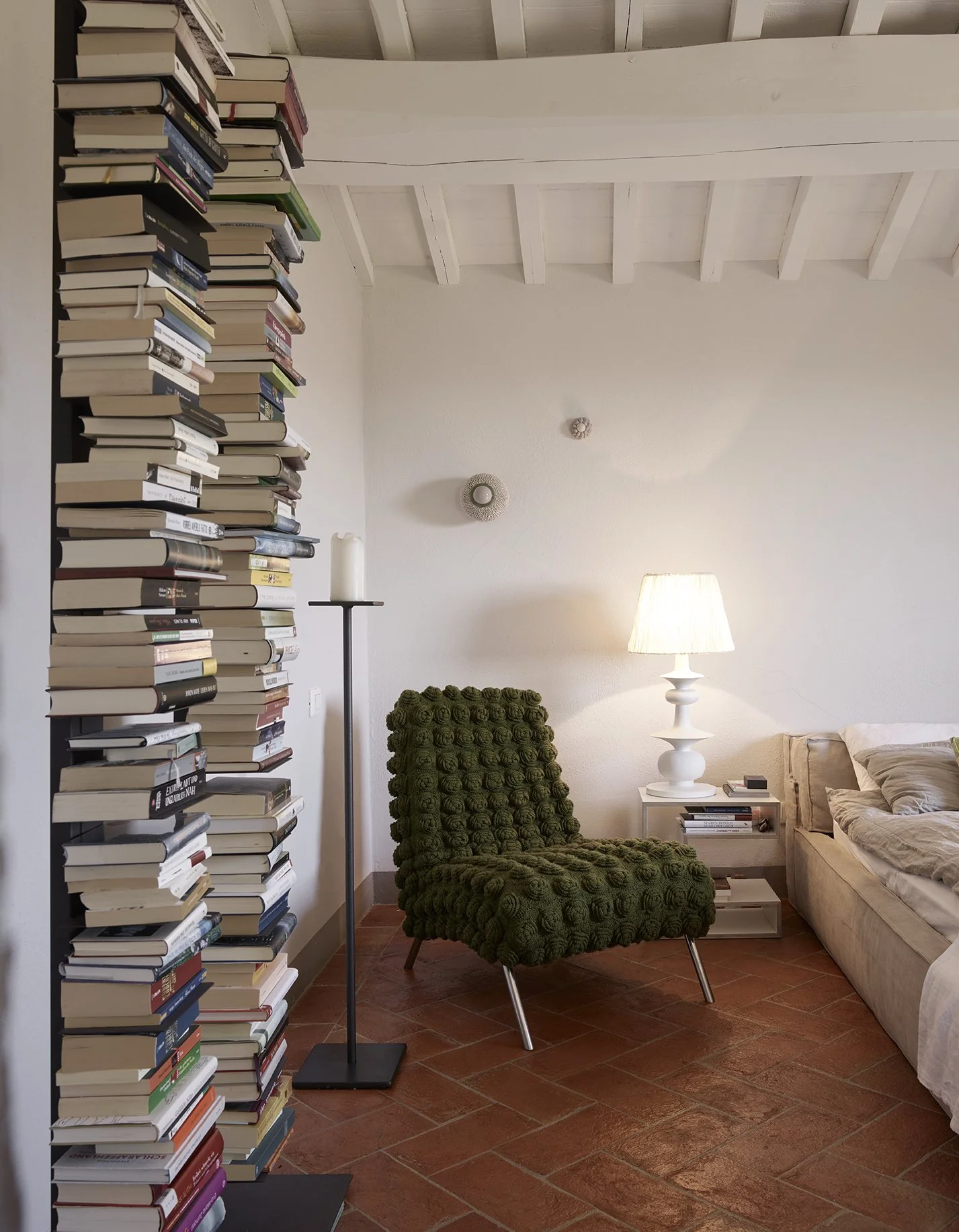 Ein Wohnzimmer mit einer hohen Bücherwand, einem grünen gepolsterten Sessel, einem Nachttisch mit Lampe, einem Sofa und hellen Wänden unter einem Holzdecke.