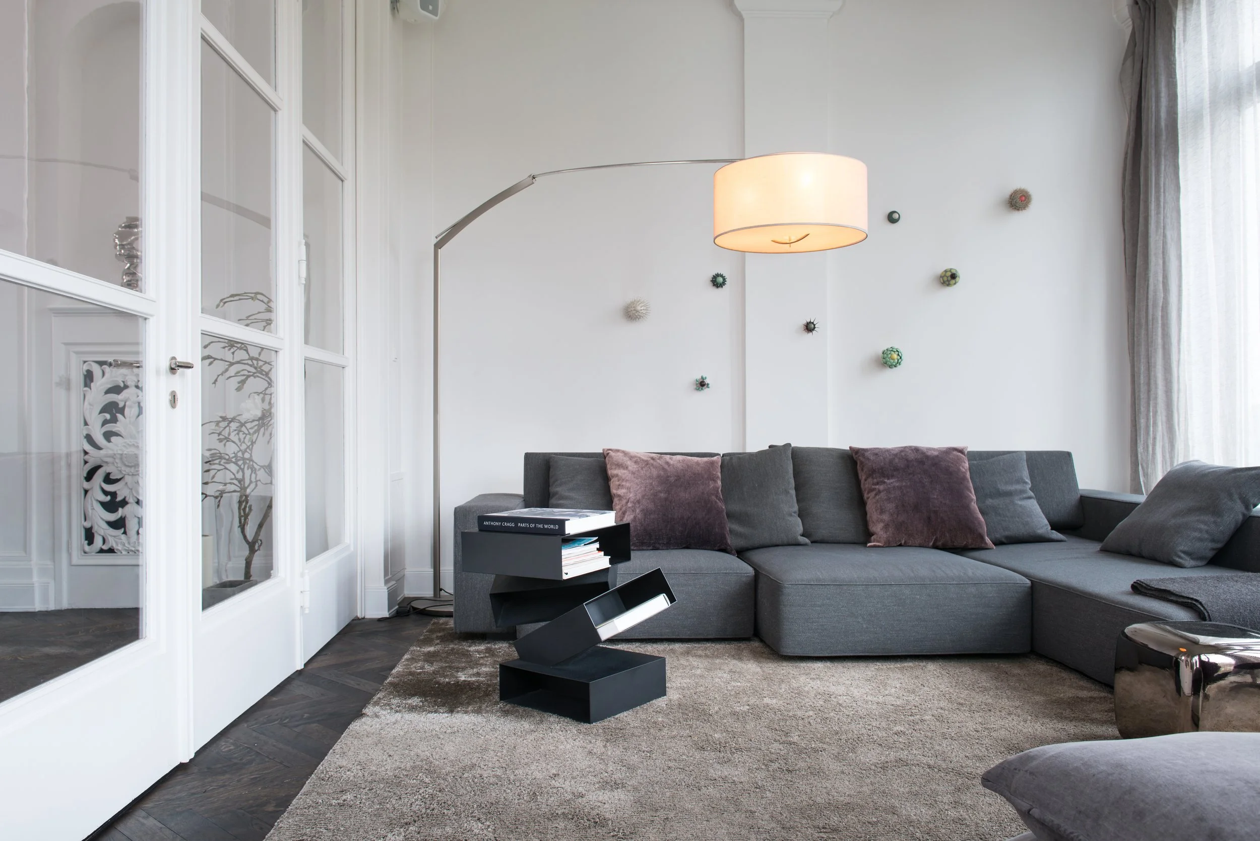 Moderner Wohnzimmer mit grauer Couch, schwarzen Beistelltisch, Teppich, Bodenkur‌ch‌en, weißen Wänden und großen Fenstern, die Vorhänge haben.