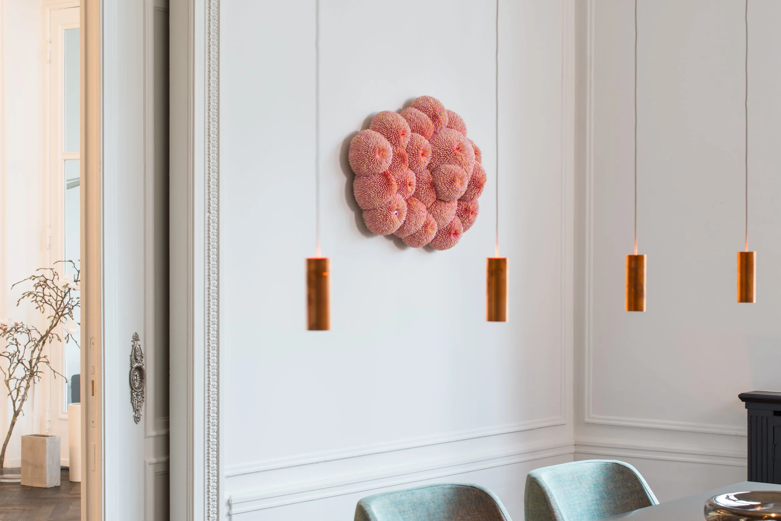 Moderne Innenraumgestaltung mit weißen Wänden, einer Wanddekoration aus rosa Muskeln, drei hängenden Kupferlampen und einem Teil eines Esszimmertisches mit Stühlen.