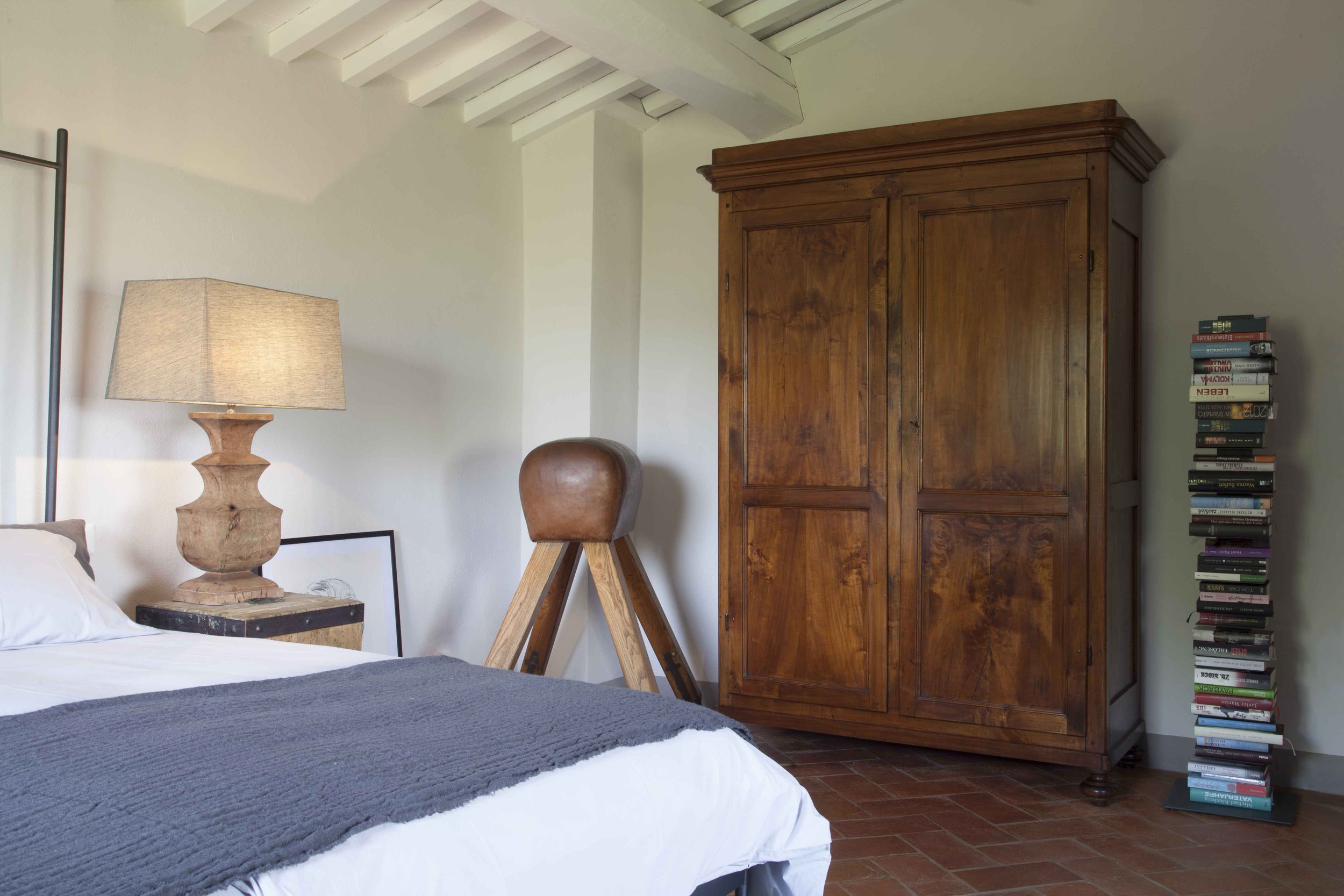 Schlafzimmer mit einem Bett, einem Nachttisch mit einer Lampe, einem Holzstuhl, einem großen Holzschrank und einem Bücherstapel neben dem Schrank.