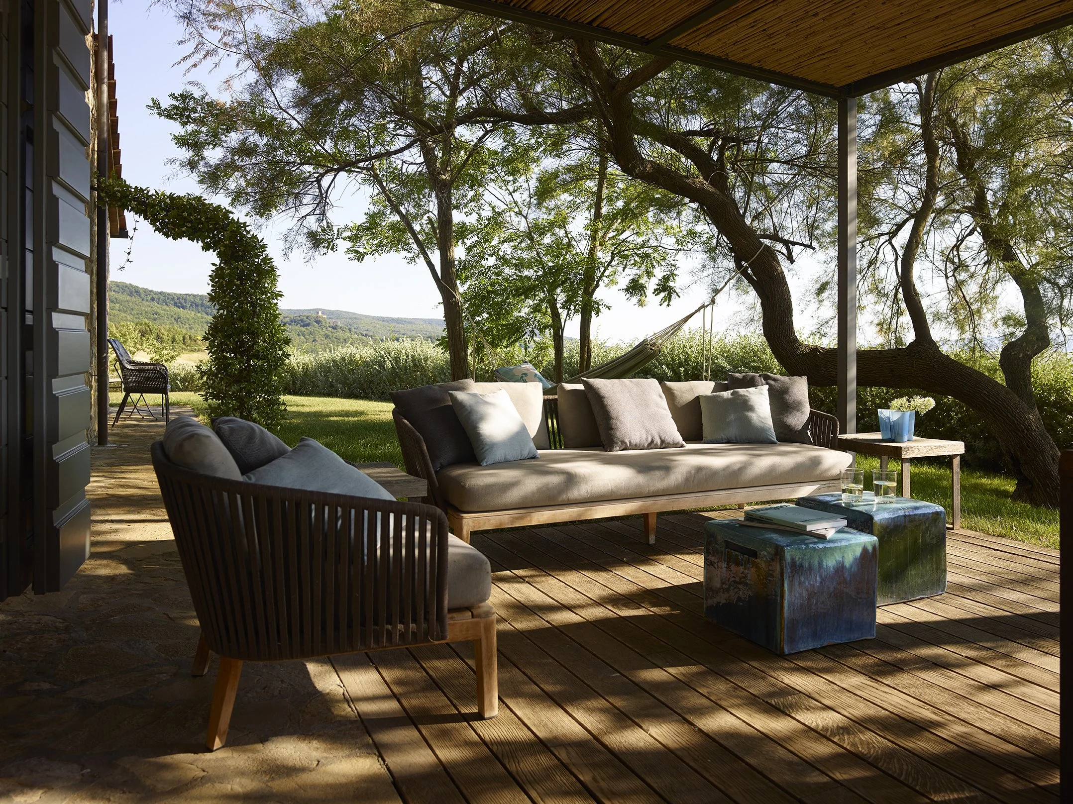 Gemütliches Outdoor-Wohnzimmer mit Spezialkissen, einem großen Sofa, einem kleinen Holztisch mit Wasser und Büchern, Sonnenlicht, Bäumen und einem Blick auf die Landschaft im Hintergrund.