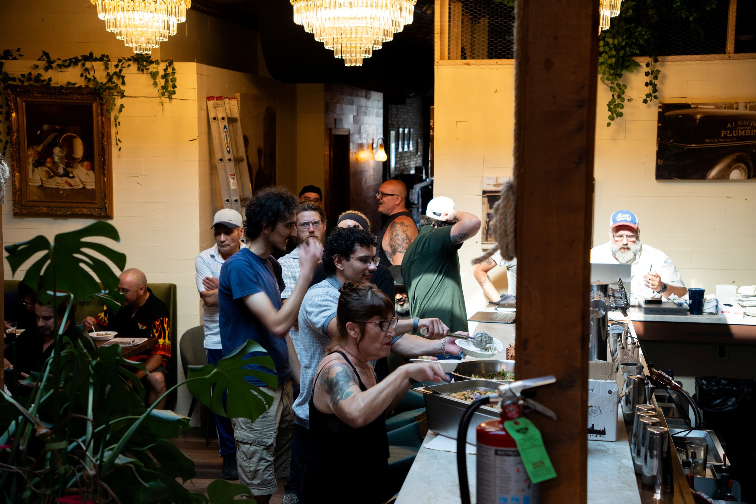 People gathered in a restaurant or cafe with warm lighting, chandeliers, and greenery, eating and socializing.