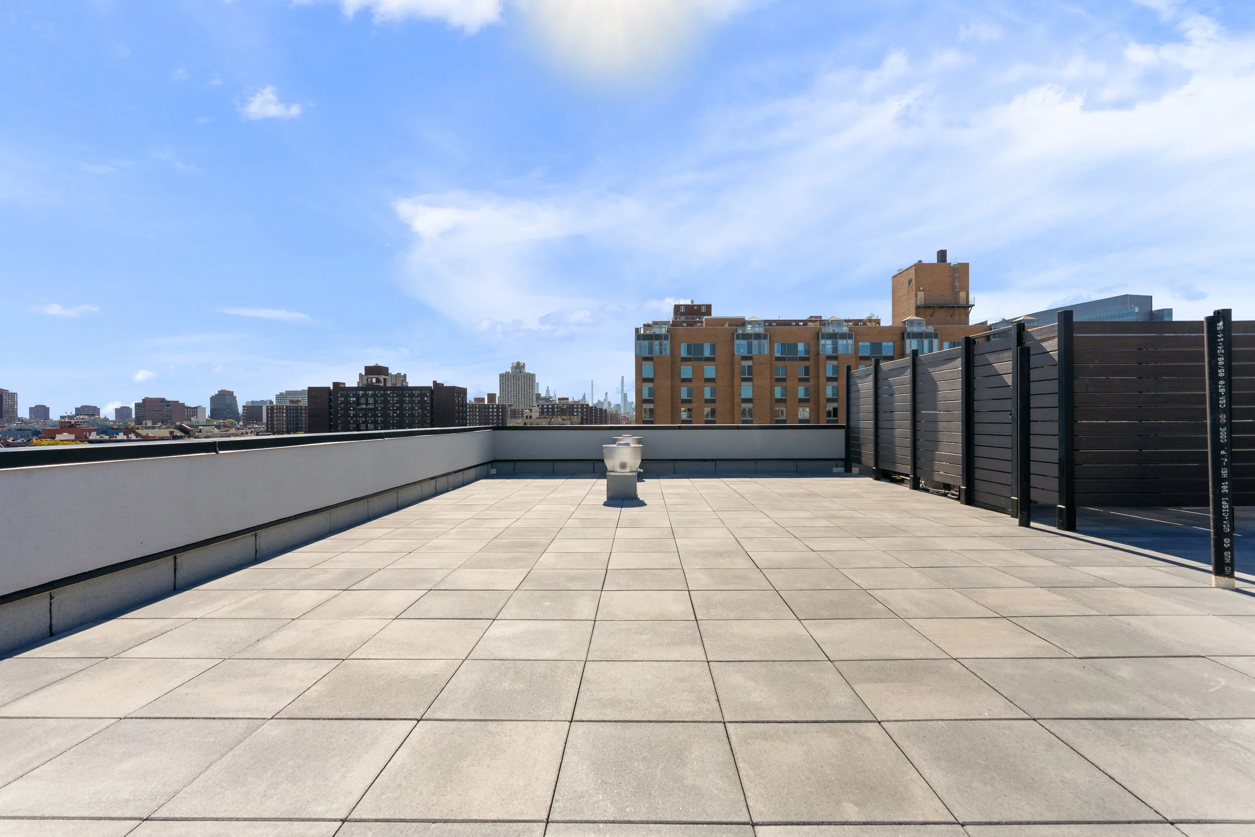 Rooftop terrace with modern beige sofas, wooden chairs, and green potted plants under a blue sky with scattered clouds, overlooking the city skyline.