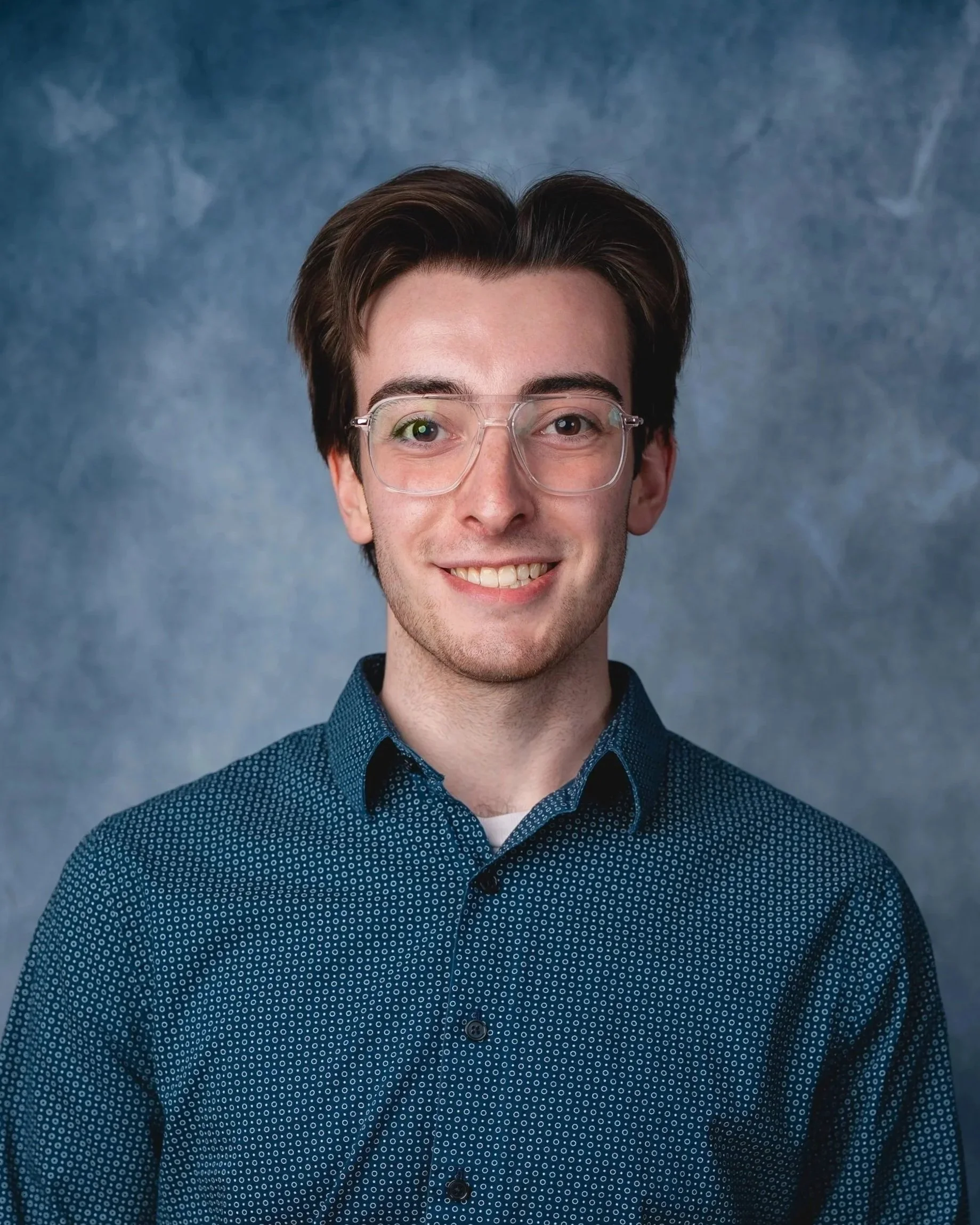 A young man with brown hair, wearing glasses and a navy blue shirt with small white dots, smiles at the camera against a mottled blue-gray background.