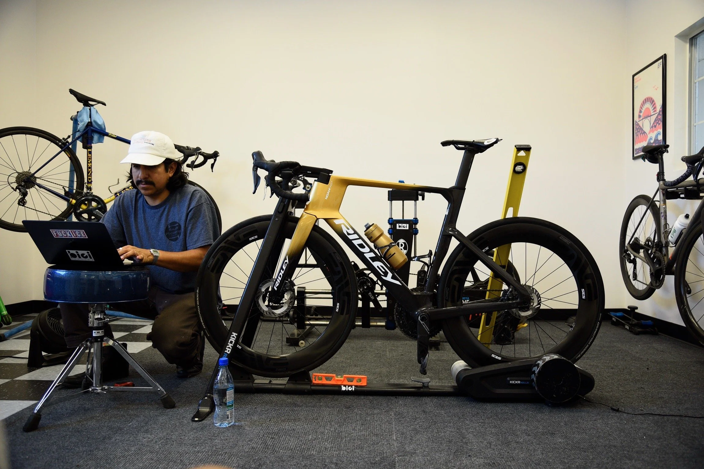 A man wearing a white hat and gray shirt sitting at a workstation with a computer, surrounded by multiple bicycles and bike maintenance equipment inside a room with black and white checkered flooring.