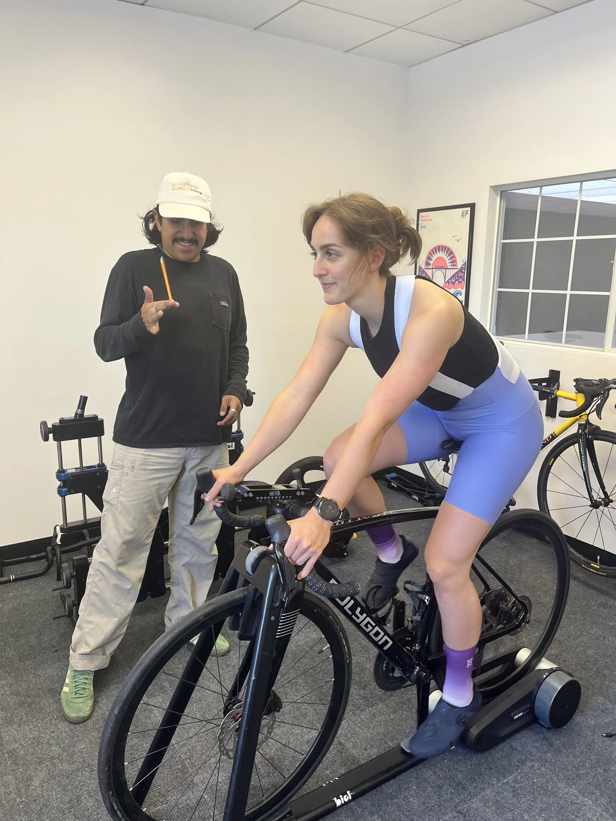 A woman in sporty attire riding a stationary bike indoors, while a man wearing a cap and casual clothes stands next to her, holding a pencil and gesturing with his hand.