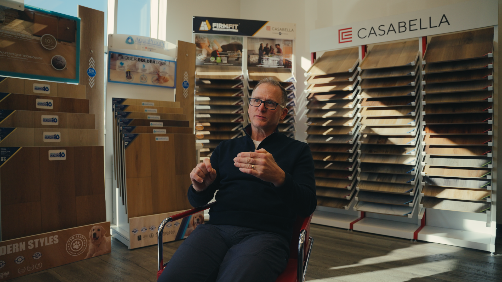 A man sitting in a store surrounded by multiple display racks of wooden flooring samples, with brand signs 'FIRM FIT' and 'CASABELLA' behind him.