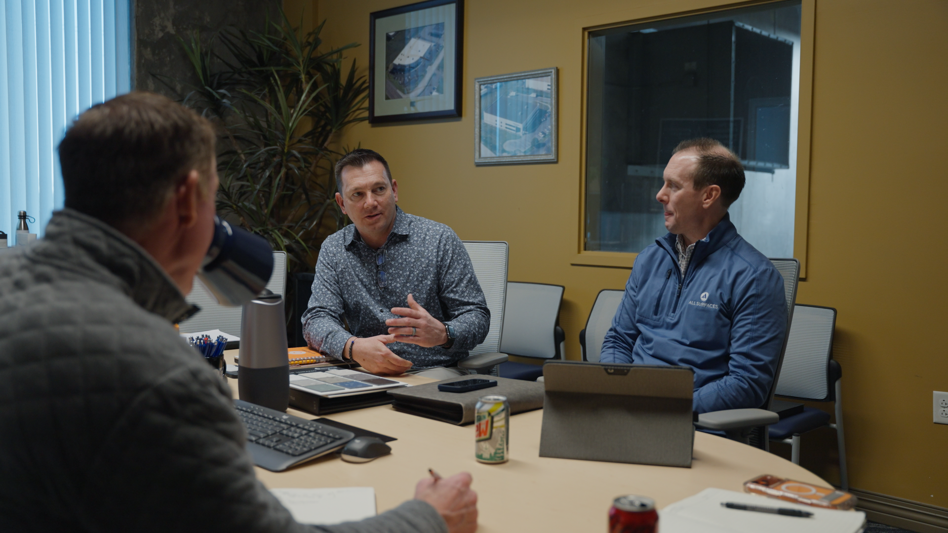 Three men are sitting around a conference table in a meeting room, engaged in discussion. The table has notebooks, a laptop, and beverage cans. There are framed pictures on the yellow wall and a large window with blinds in the background.