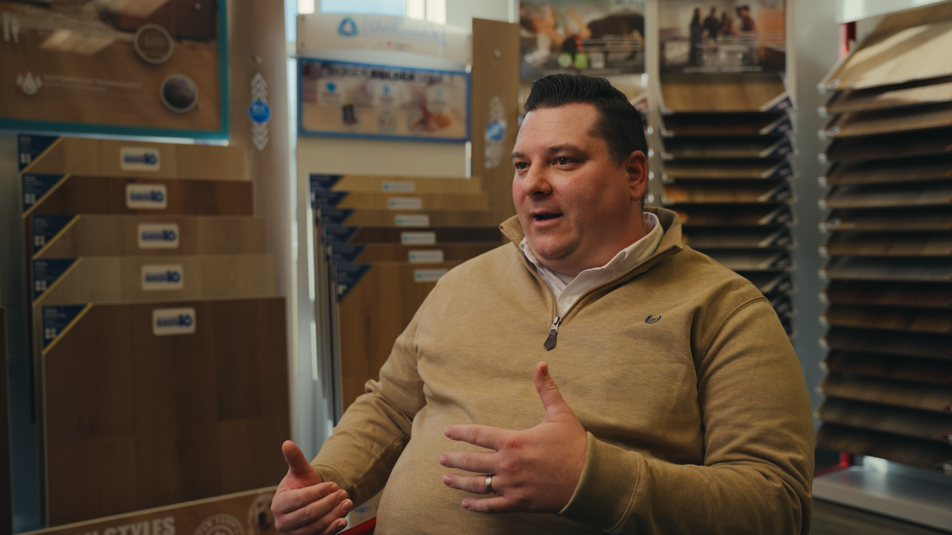 A man with dark hair, wearing a tan zip-up sweater over a white shirt, is sitting in a furniture store surrounded by wood flooring samples on display racks. He appears to be engaged in conversation, with his hands gesturing.