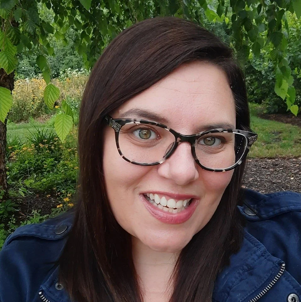 A woman with dark brown hair, wearing glasses with a tortoise shell pattern, smiling outdoors with green trees and plants in the background.