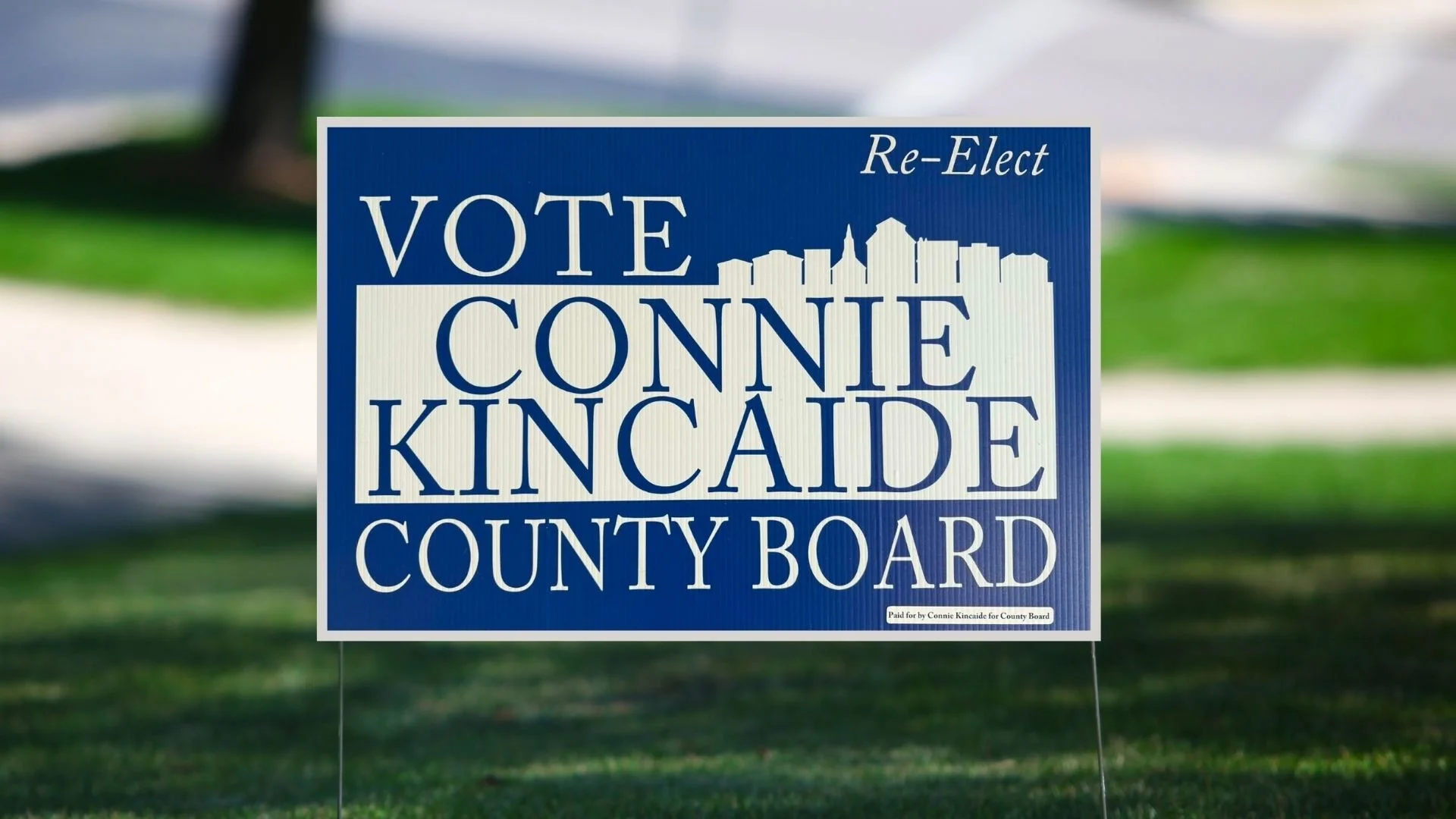 A political campaign yard sign with a blue background and white text that reads 'Vote Conne Kincaide County Board,' with a city skyline graphic and the words 'Re-Elect' in the top right corner.