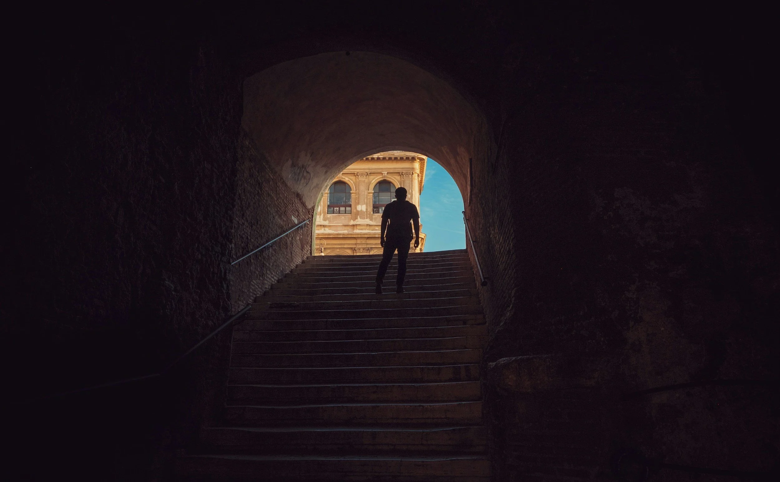 Person walking up stairs through a shadowy tunnel toward a bright, historic building with a clear blue sky in the background.