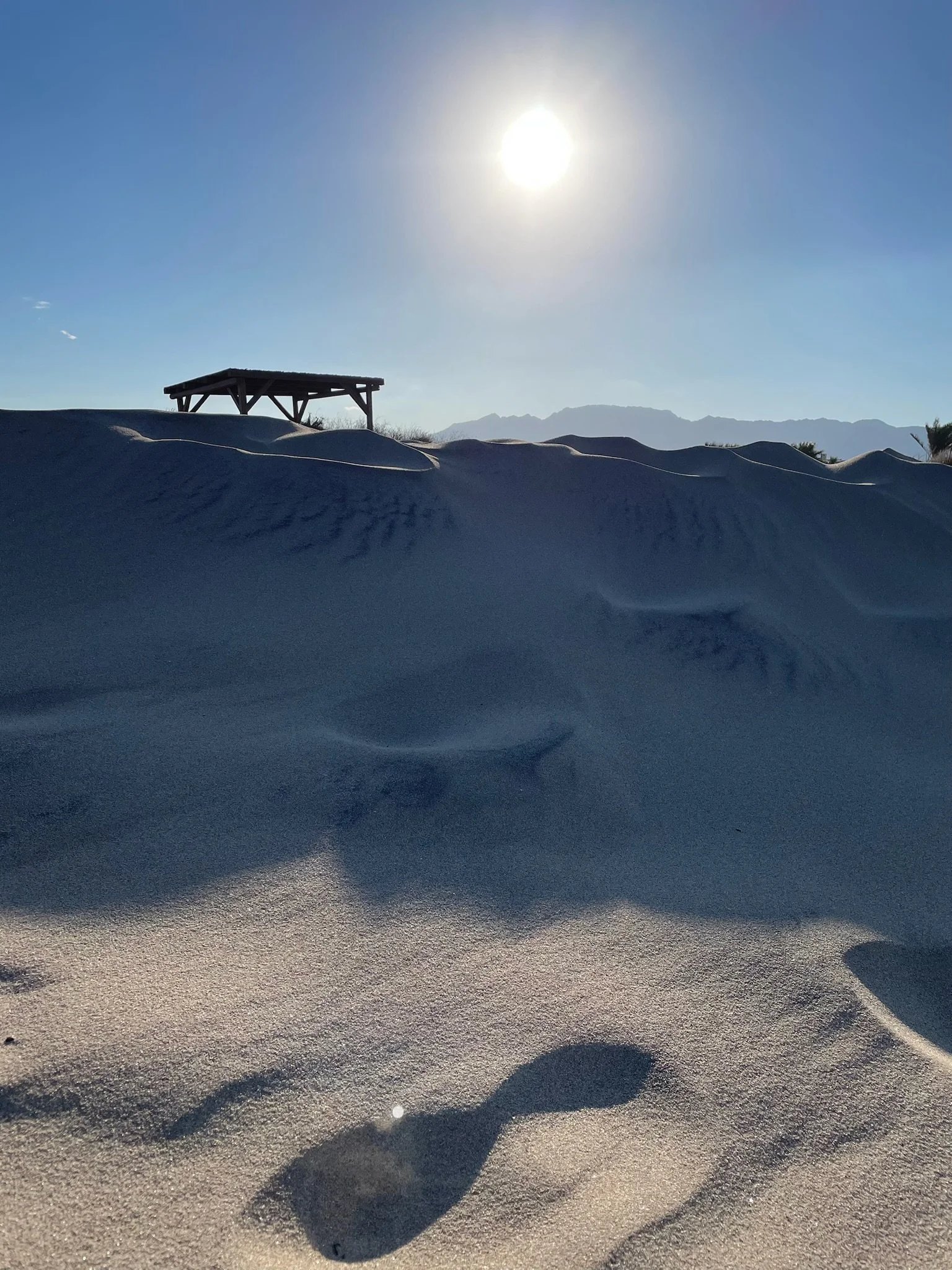 A bright sun over sand dunes with a small wooden shelter in the distance and mountains on the horizon.