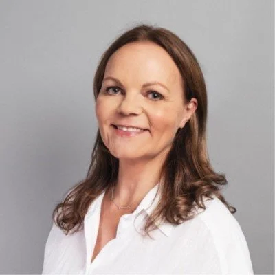 A woman with long brown hair wearing a white shirt, smiling against a gray background.