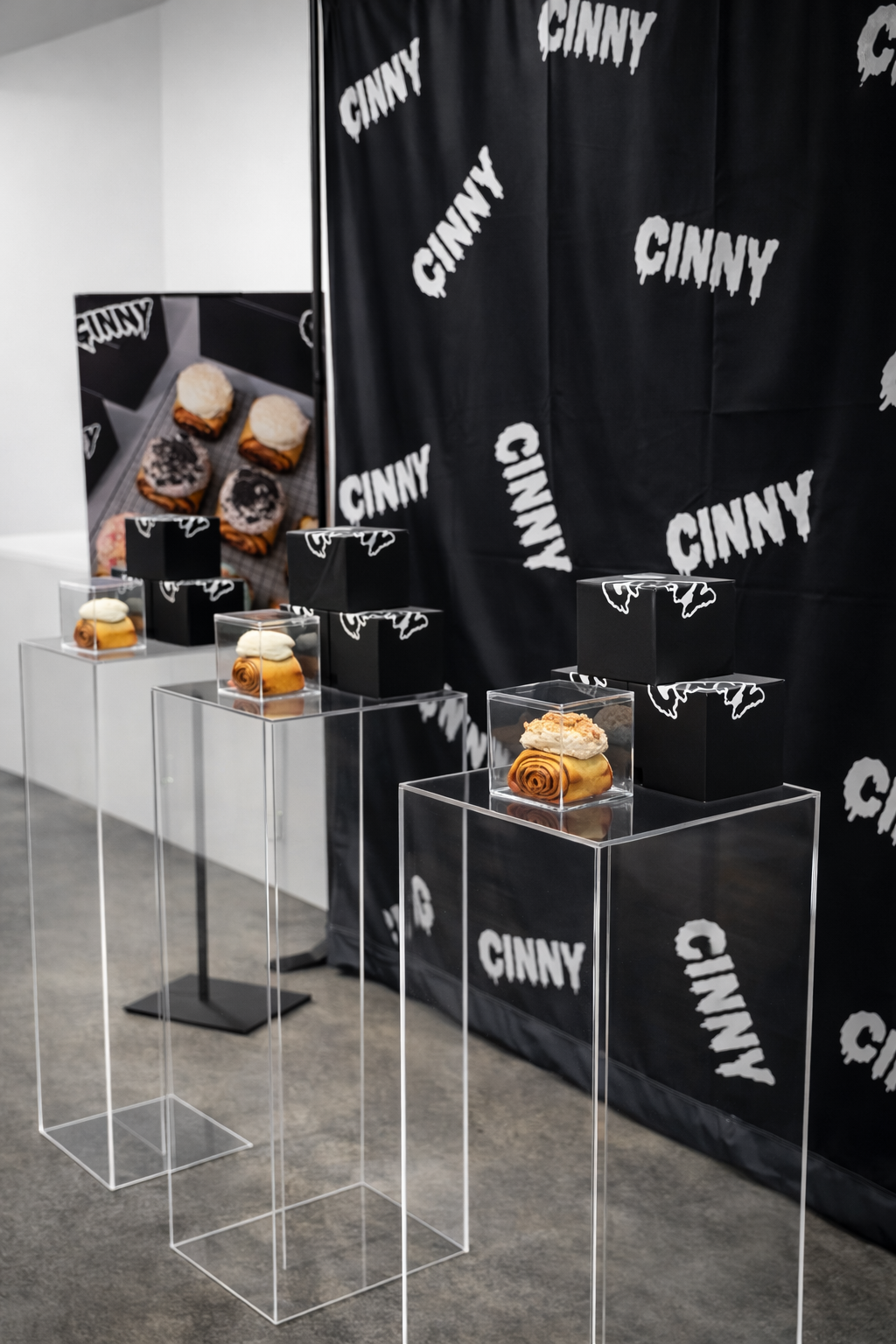 Display of 3 cinnamon rolls on transparent pedestals, with a black backdrop and promotional poster featuring cinnamon rolls and the Cinny logo.