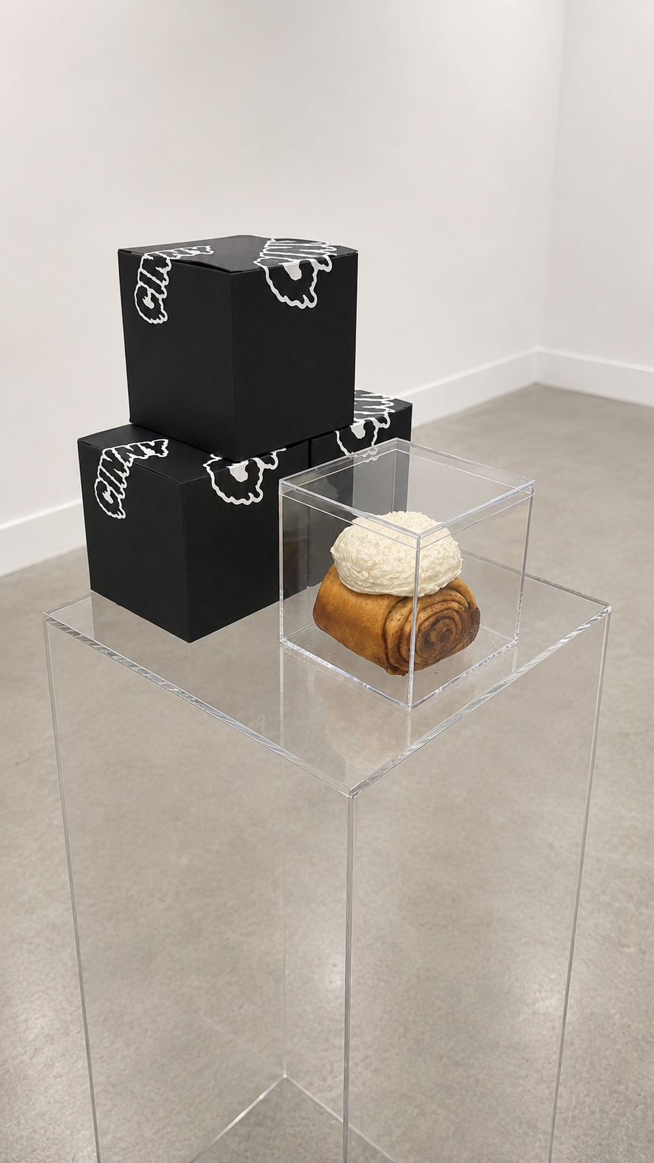 A display featuring a cinnamon roll topped with white icing inside a clear acrylic box, with three black boxes on top of each other.