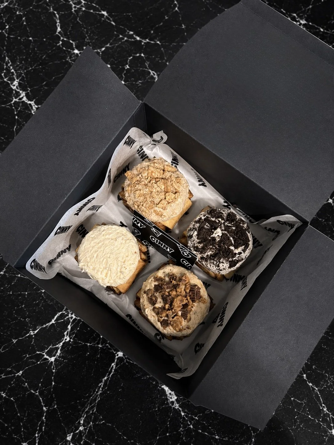 Inside a black box, four cinnamon rolls topped with a variety of frostings placed on a marble surface.