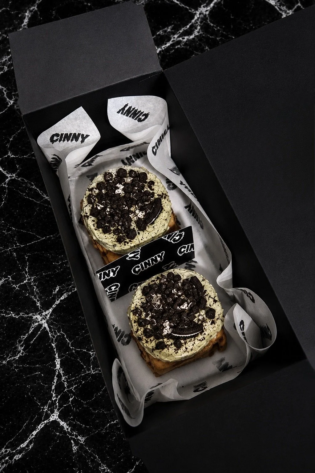 Inside a black box, two cinnamon rolls topped with green matcha frosting placed on a marble surface.