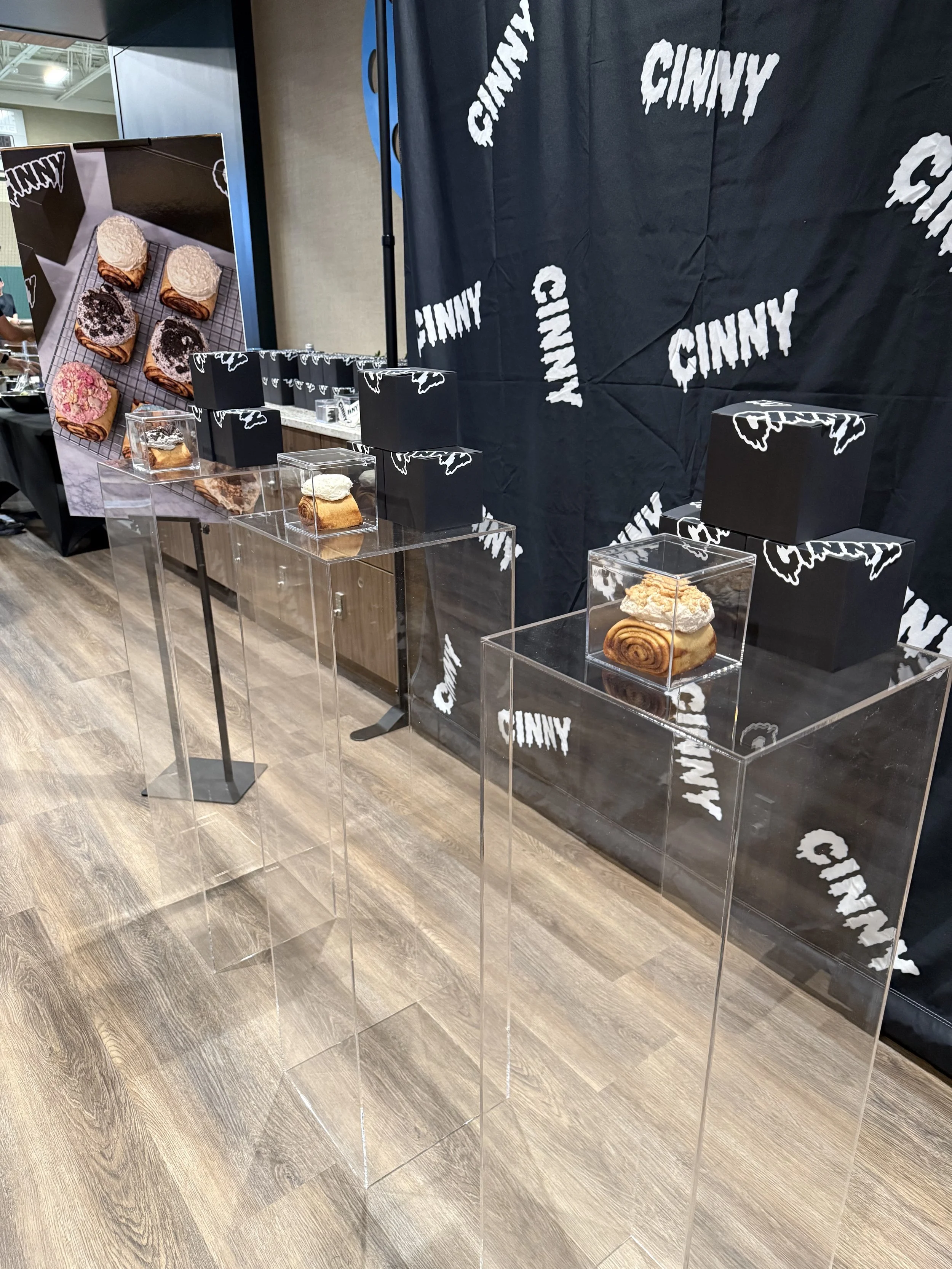 A group of cinnamon rolls topped with frosting in a clear display case on an acrylic pedestal.