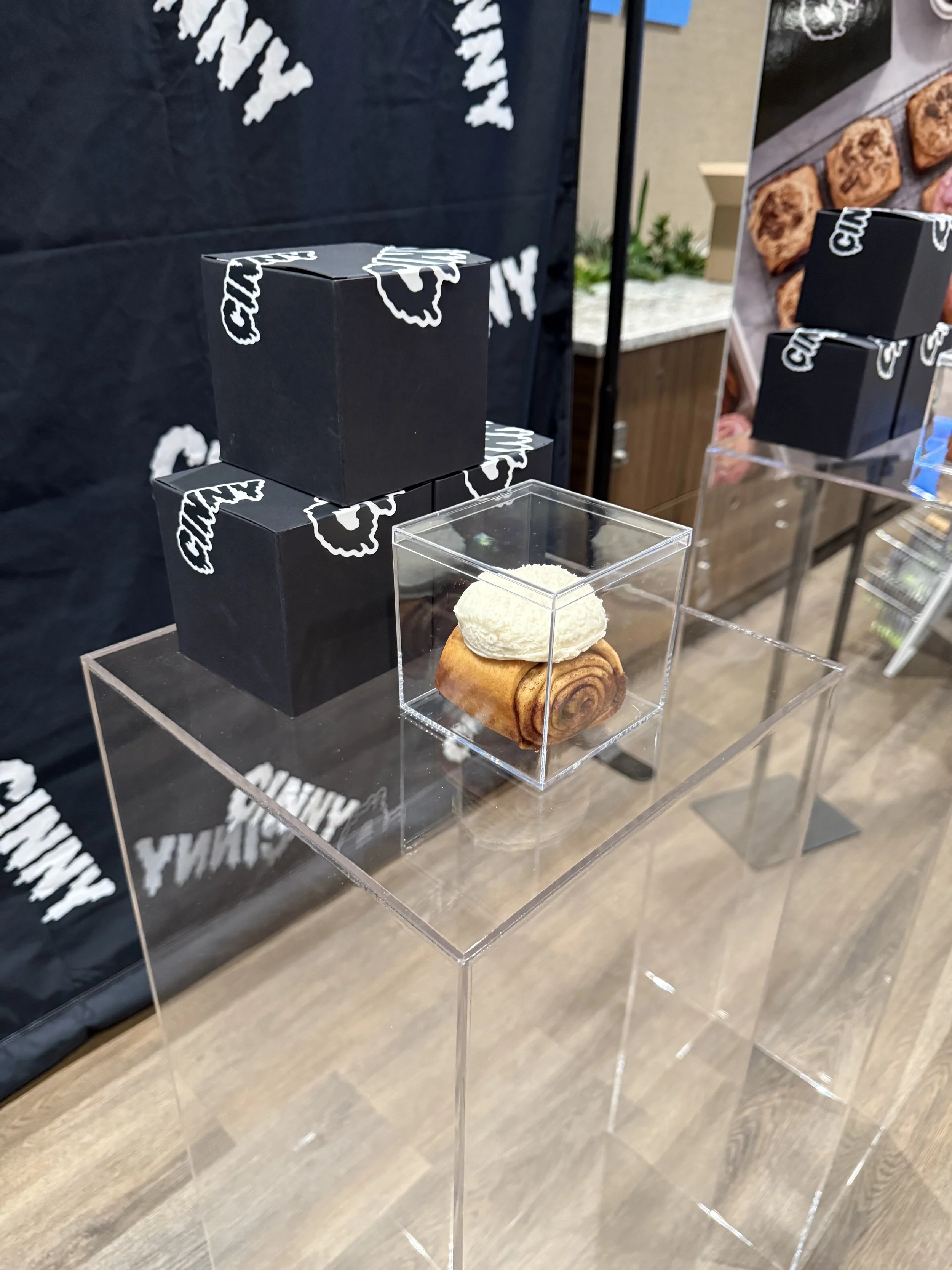 Display of a cinnamon roll with a scoop of vanilla ice cream on top, enclosed in a transparent acrylic box, with black boxes and posters in the background at a bakery or food event.