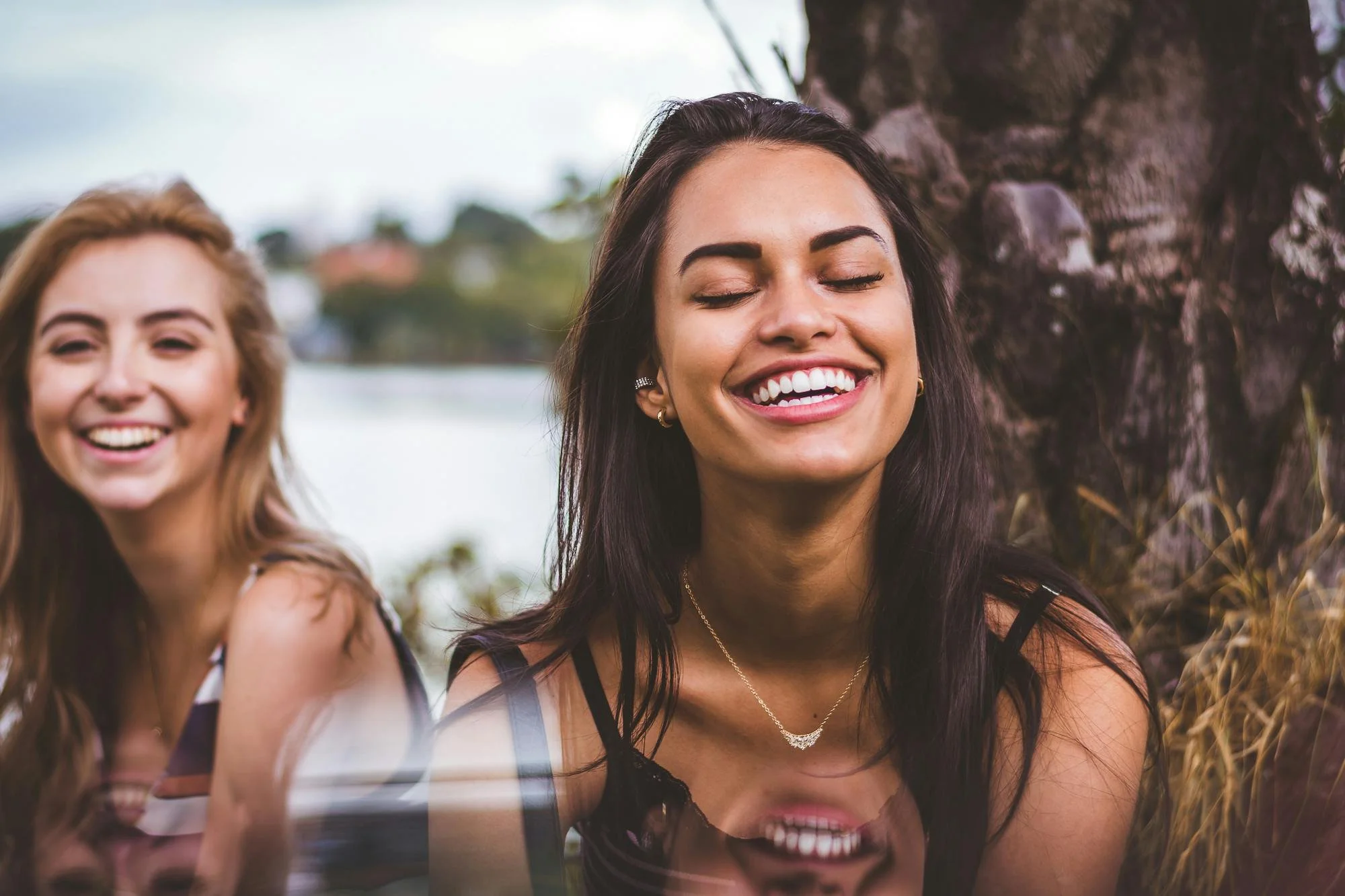 Two young women smiling outdoors near a tree with water in the background.