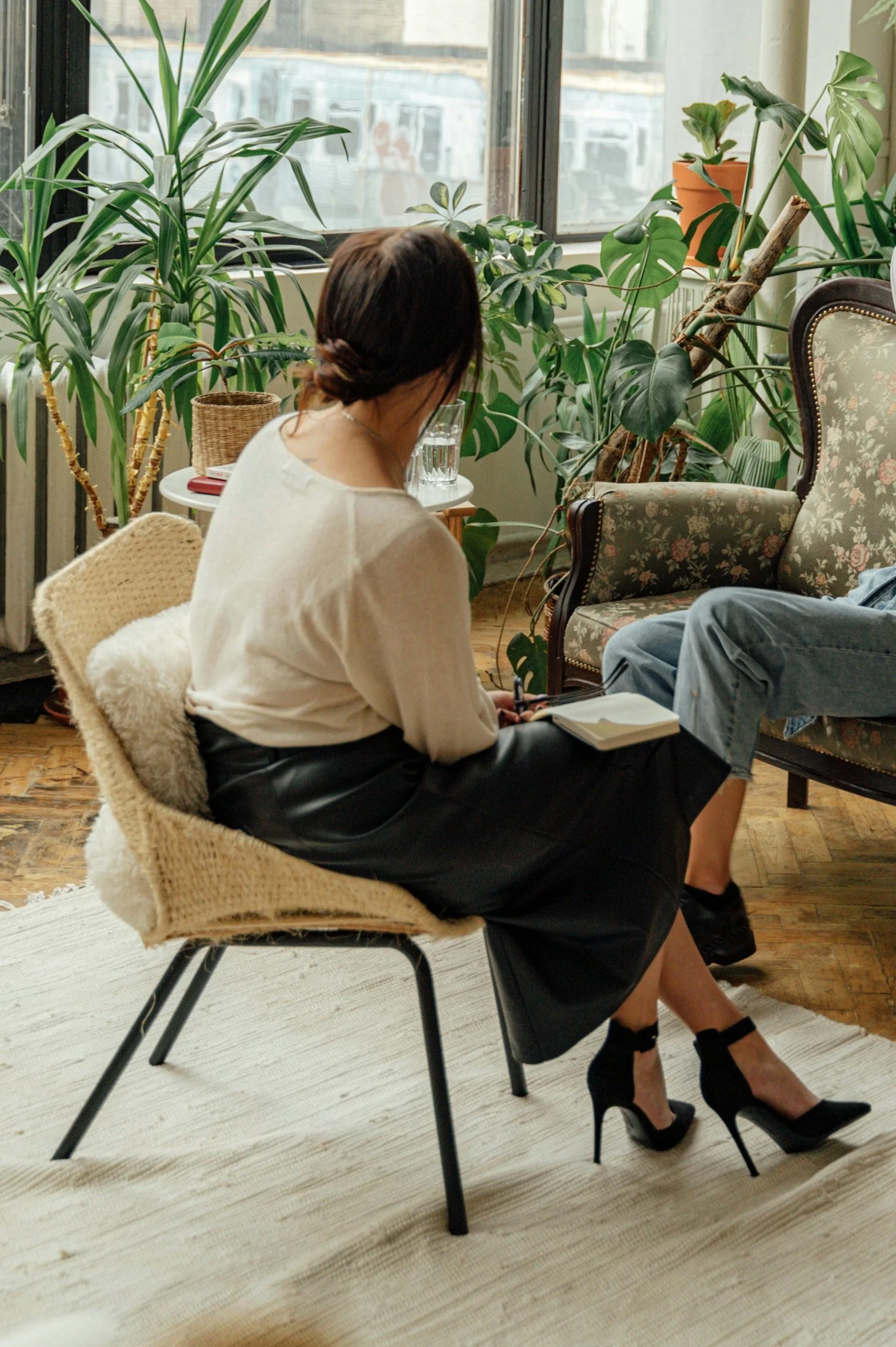 A woman sitting with her back to the camera, taking notes in a room filled with green plants, a floral armchair, and a large window.