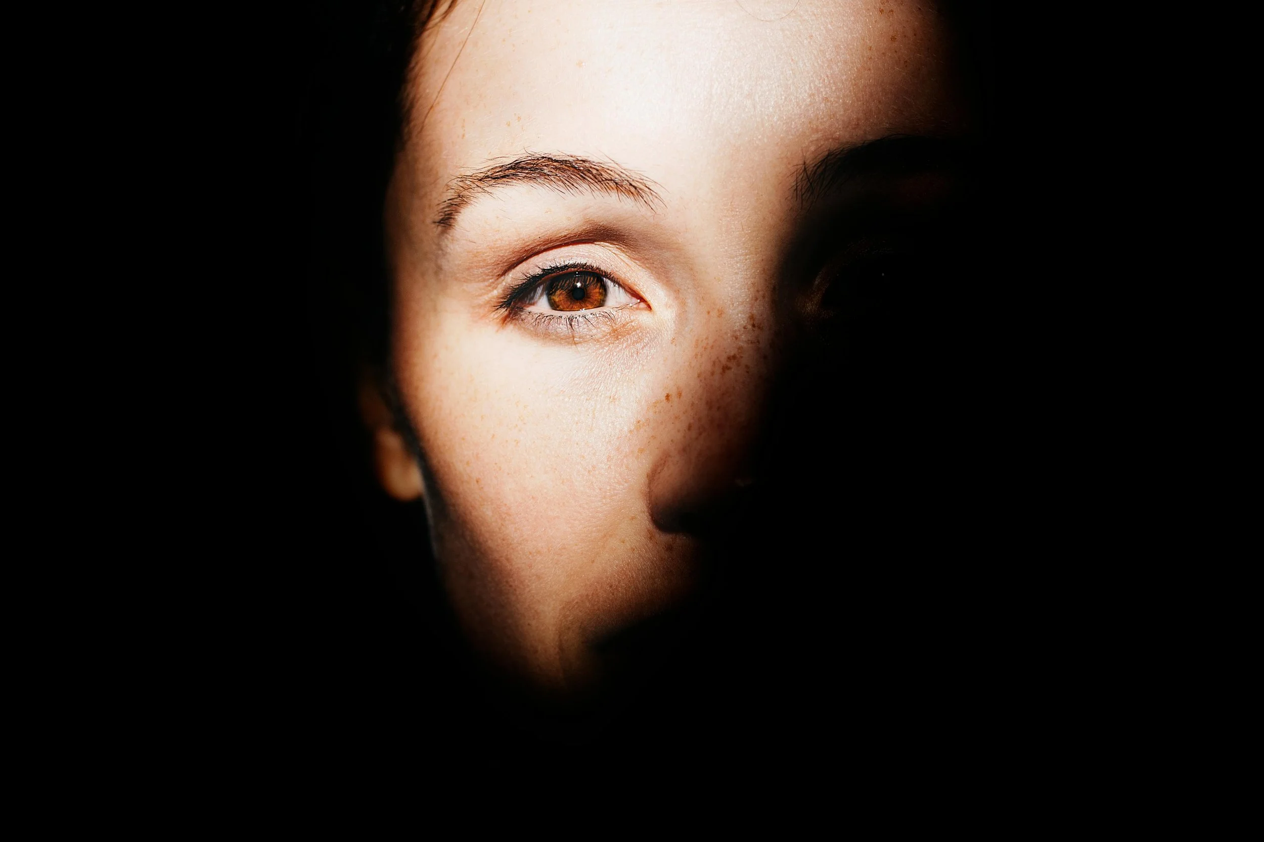 Close-up of a young woman's face, partially illuminated, with brown eyes, freckles, and dark hair, surrounded by darkness.