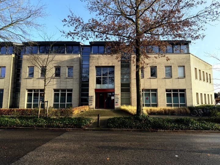 Modern multi-story residential or office building with beige brick exterior, large windows, and a central entrance, surrounded by trees and landscaping.