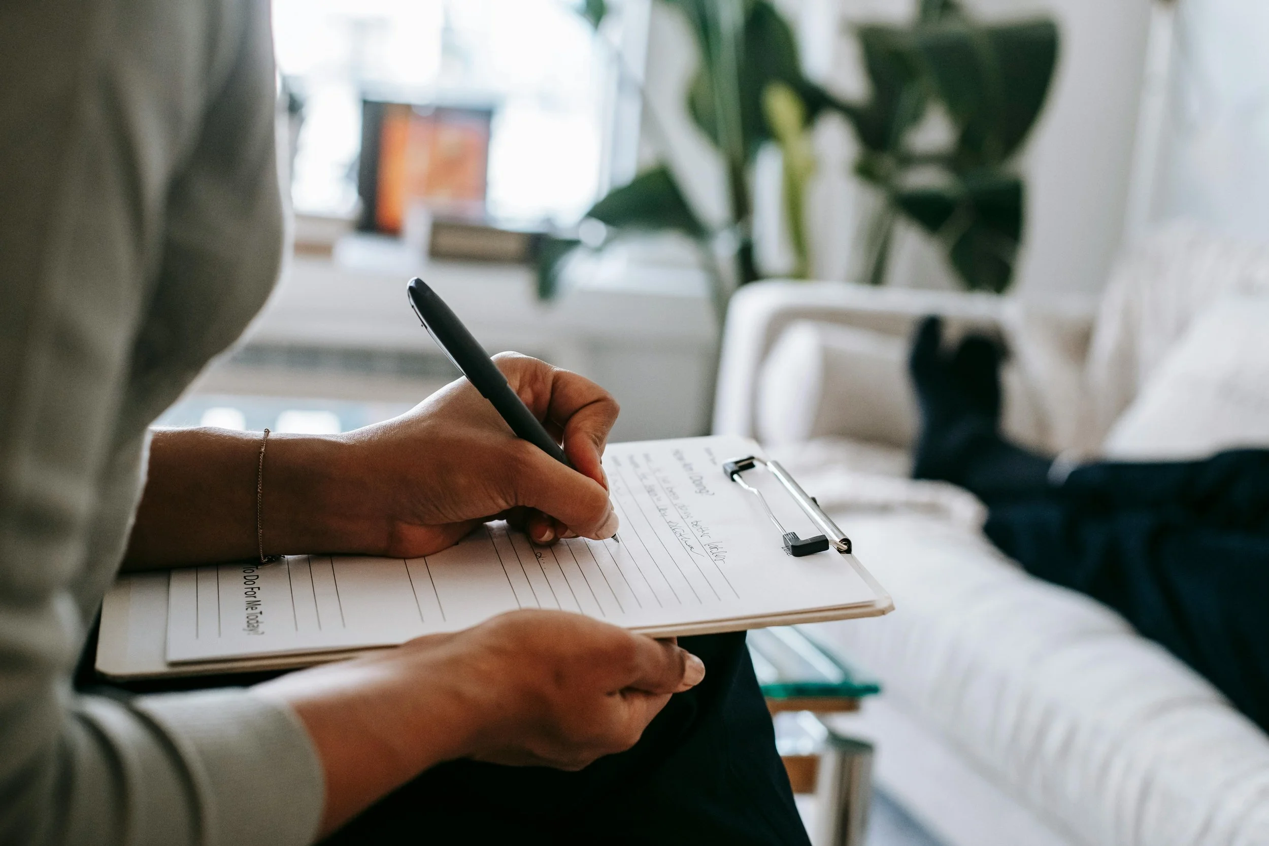 Person taking notes on a clipboard while lying on a couch in a well-lit living room.