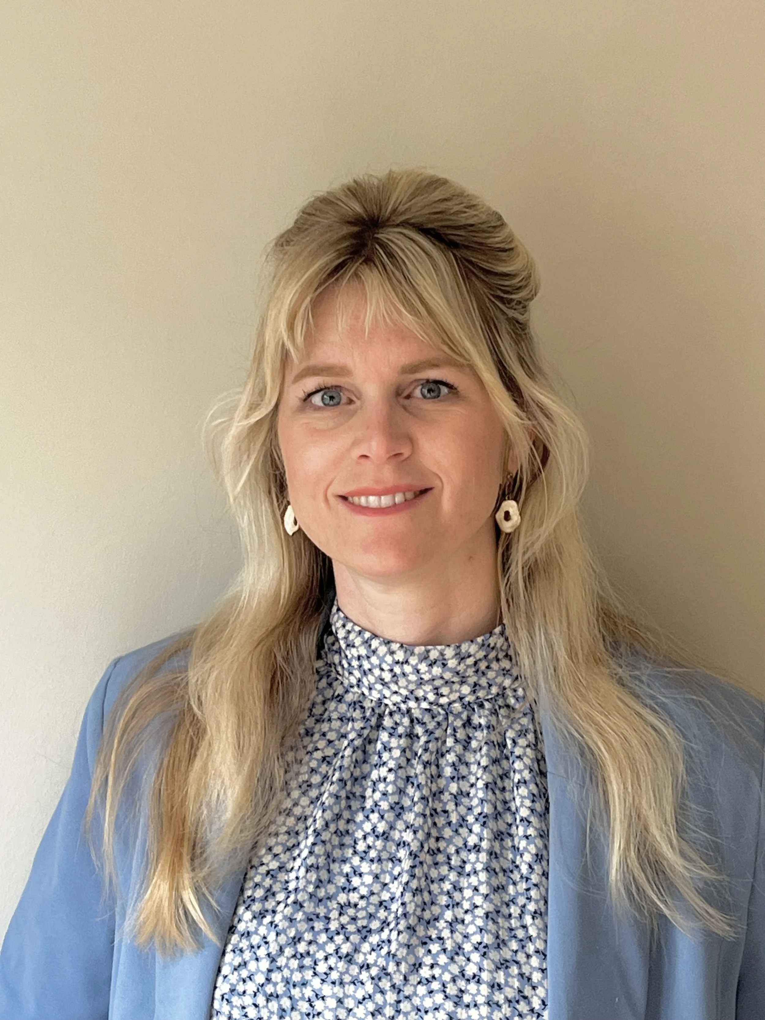 A woman with blonde hair wearing pearl earrings, a patterned blouse, and a light blue blazer standing against a plain light-colored wall.