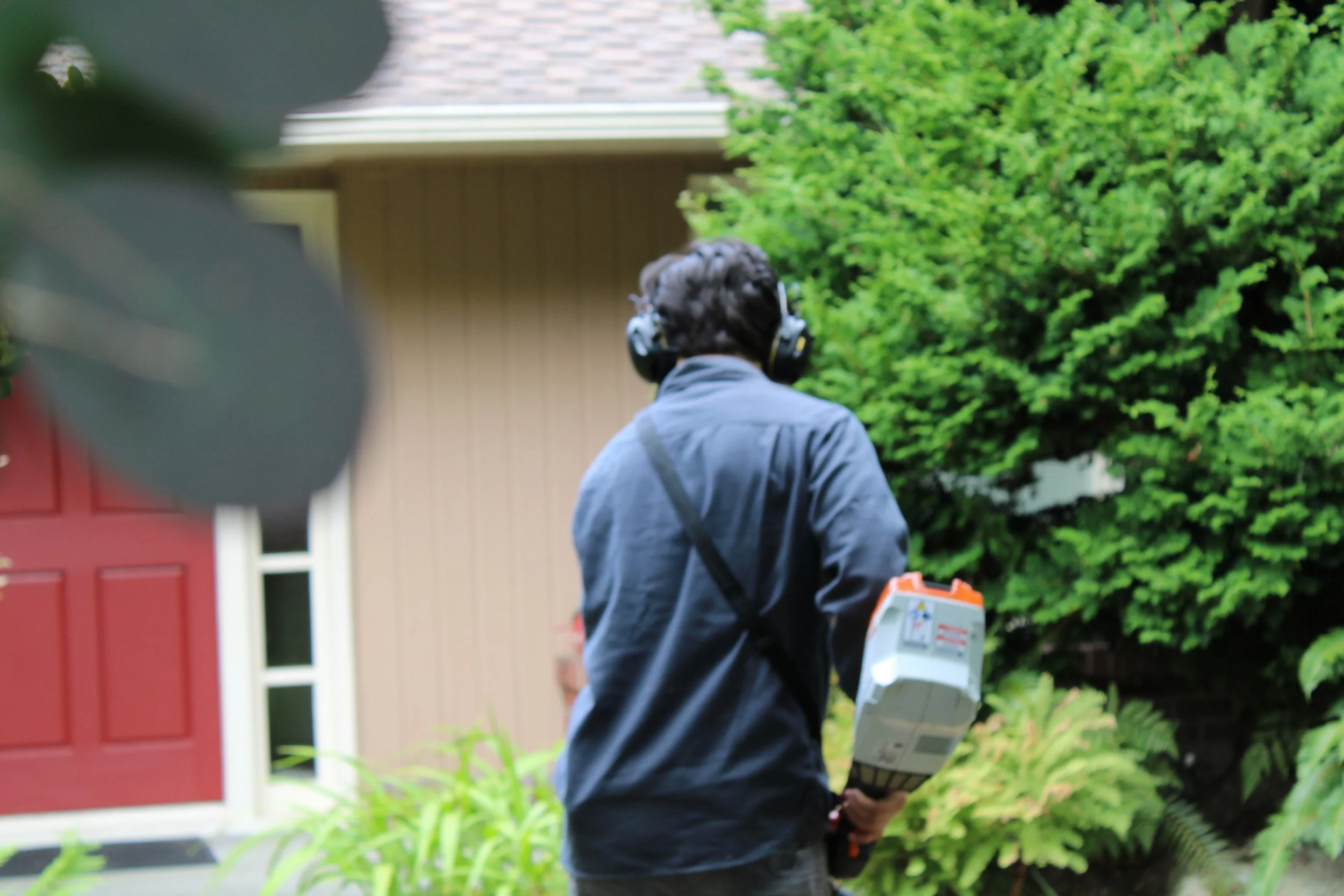 A person in blue jacket wearing noise-canceling headphones, holding a device, walking outdoors near a house with a red door and lush green trees.