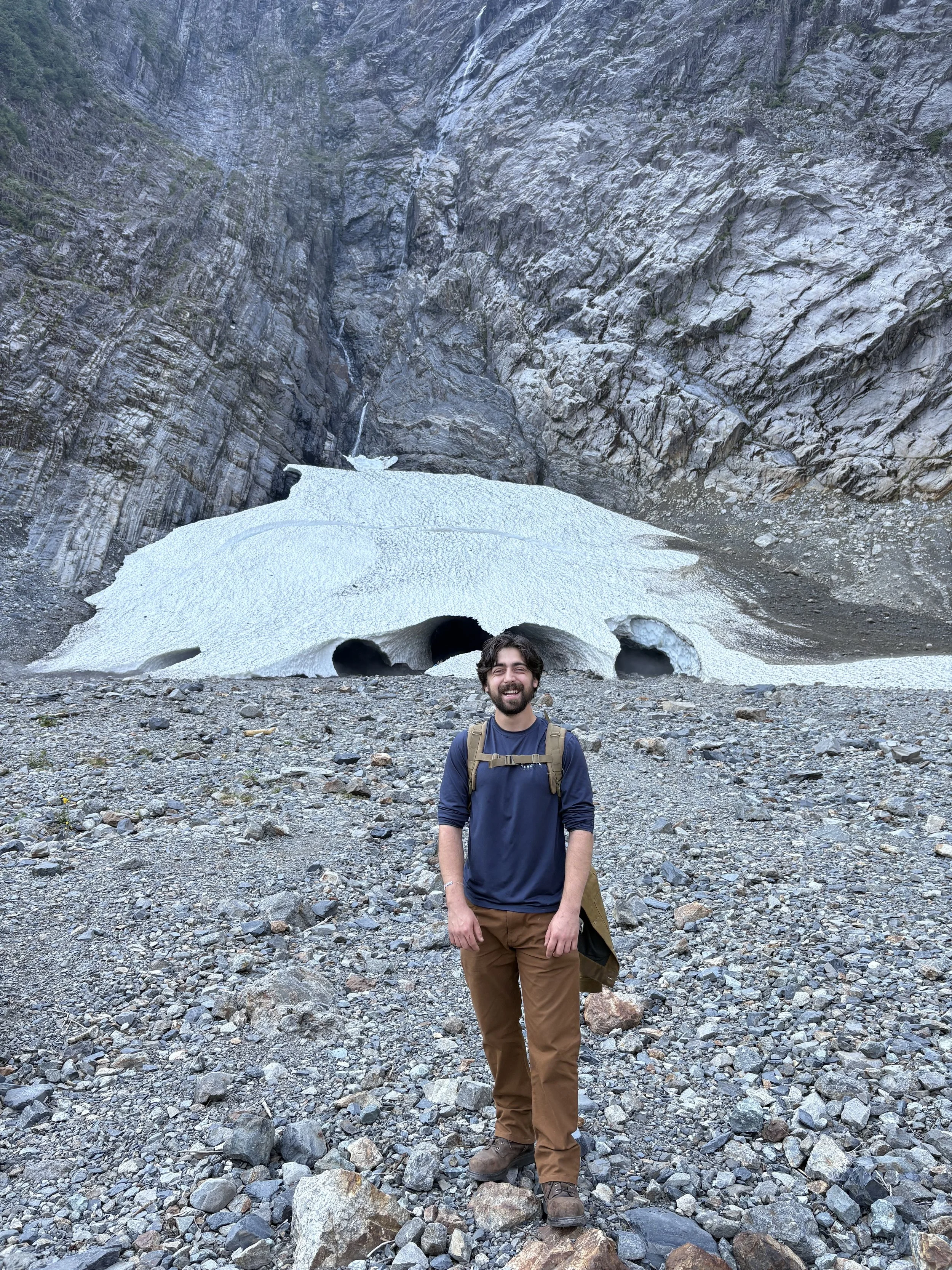A man with a backpack standing on rocky terrain in front of a glacier with small cave openings, surrounded by high mountain cliffs.