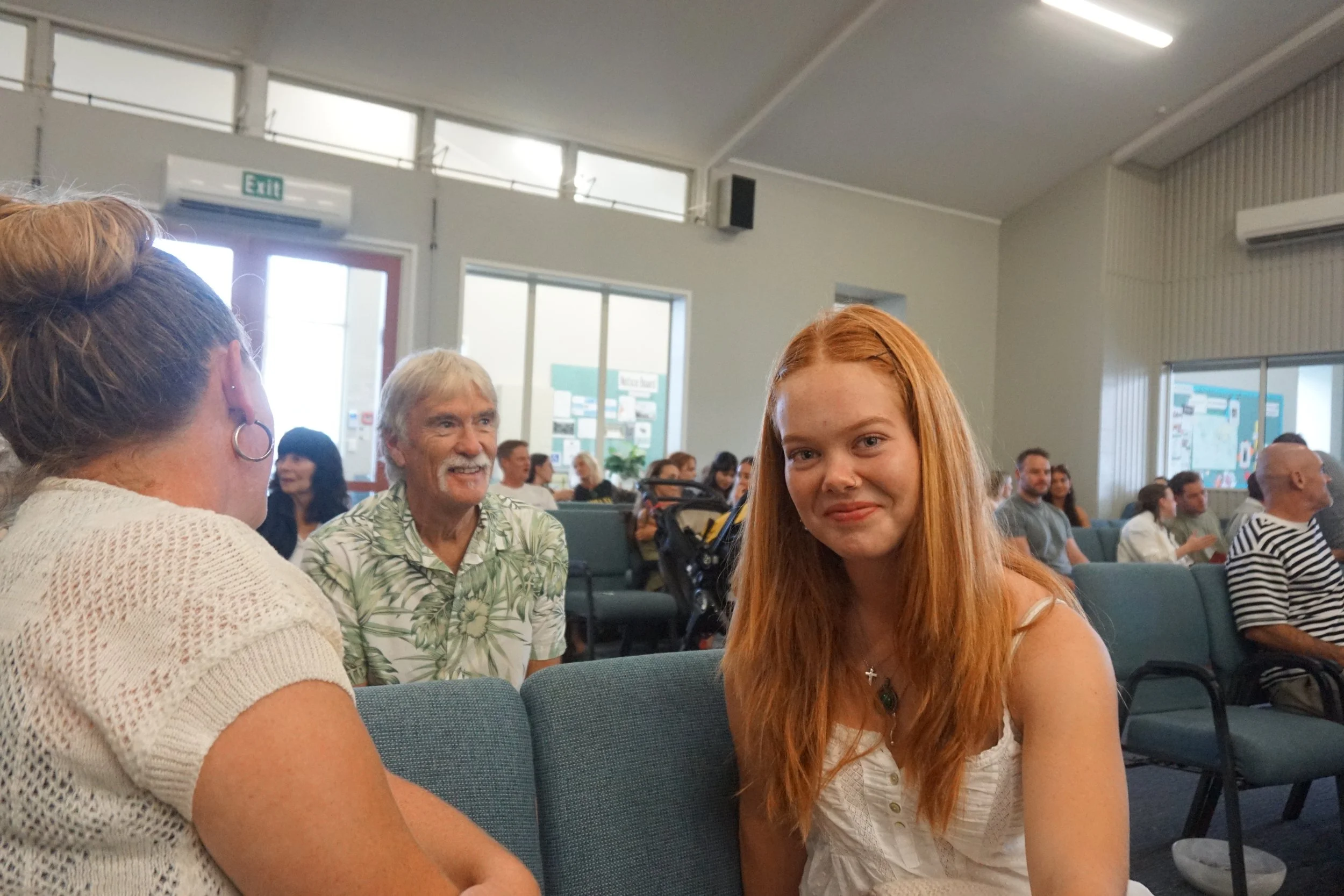 Young woman with long red hair sitting in a room, smiling at the camera, surrounded by other people.