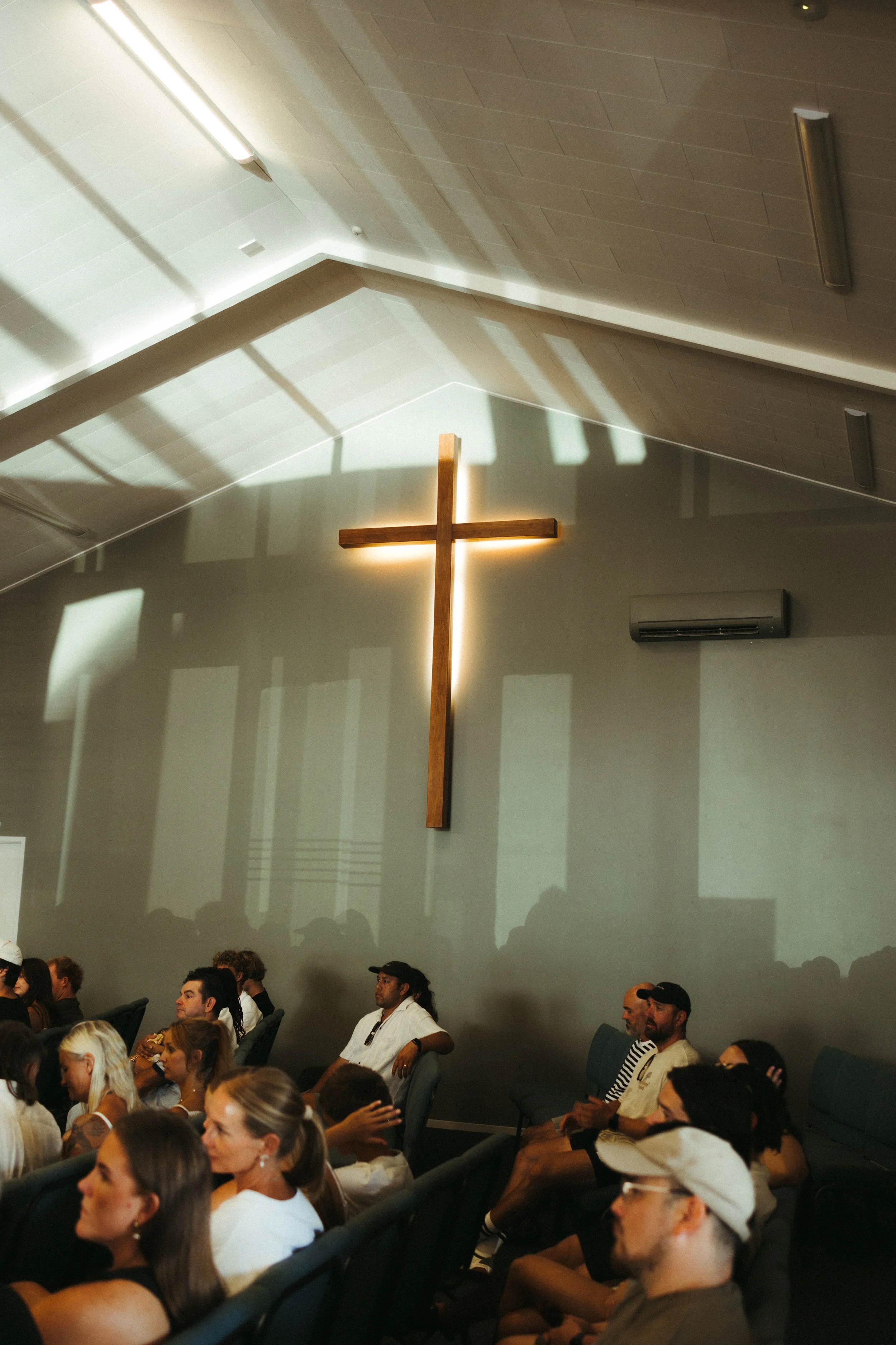 A church interior with a wooden cross illuminated on a grey wall, and people seated on chairs in the congregation.