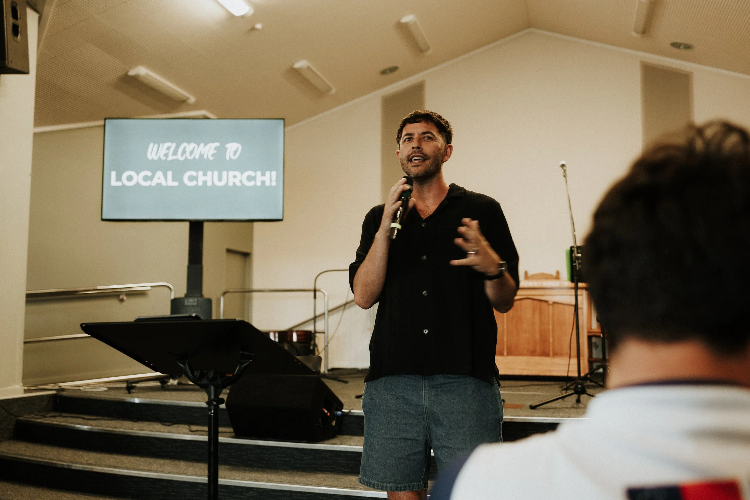 A man speaking into a microphone in front of a screen that reads 'Welcome to Local Church!' during a church service or event.