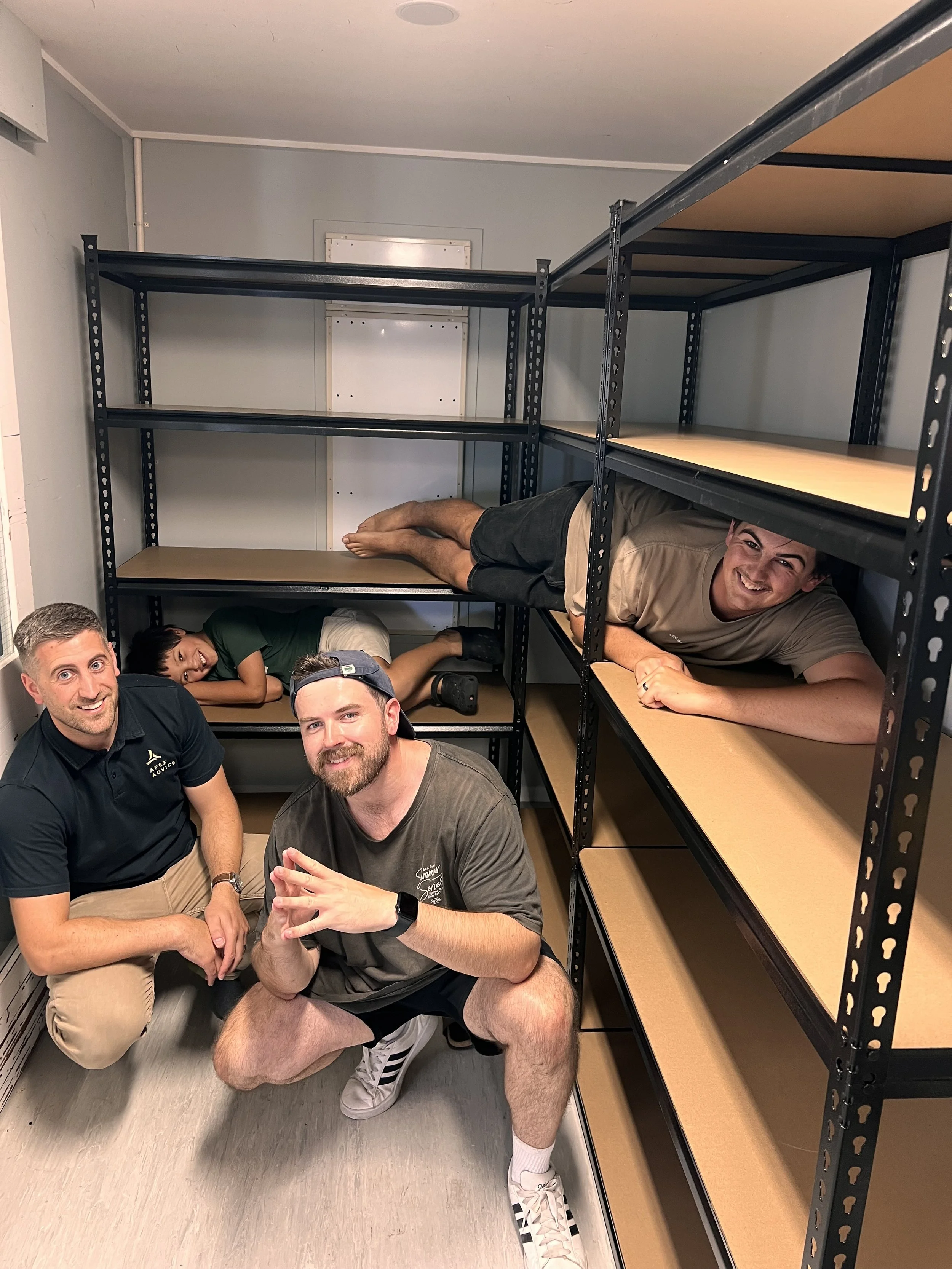 Four people are posing inside and around empty shelving units in a room with light-colored walls. One person is lying on the middle shelf, another is lying on the right shelf, and two are crouching in front of the shelving. All are smiling at the cam